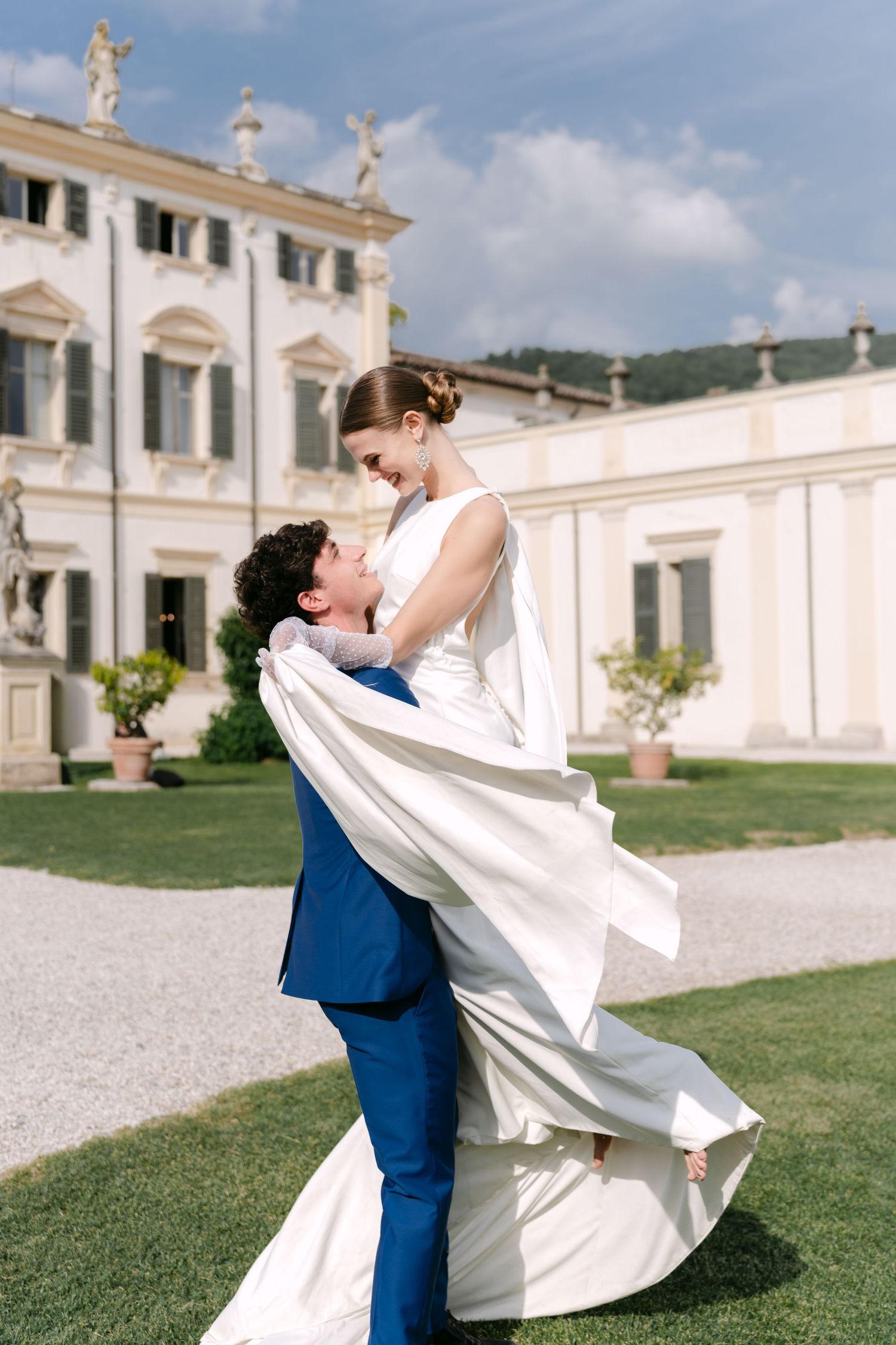 Romantic couple embraces in front of an elegant white Italian villa with classical architecture and manicured grounds. Villa Mosconi Bertani