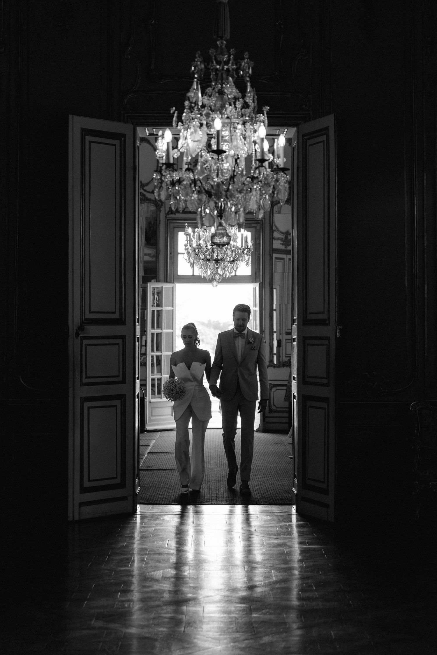 Silhouettes of two people walking through an elegant doorway with a crystal chandelier in a dark interior hallway. Schloss Solitude