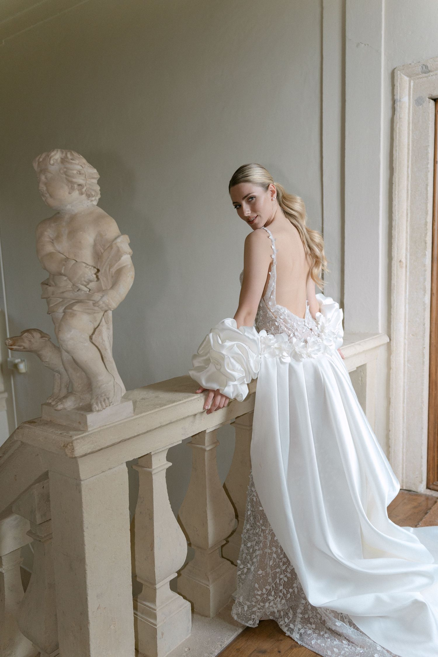 Elegant white wedding dress with open back design and flowing train shown against classic architectural balustrade. Wedding Verona