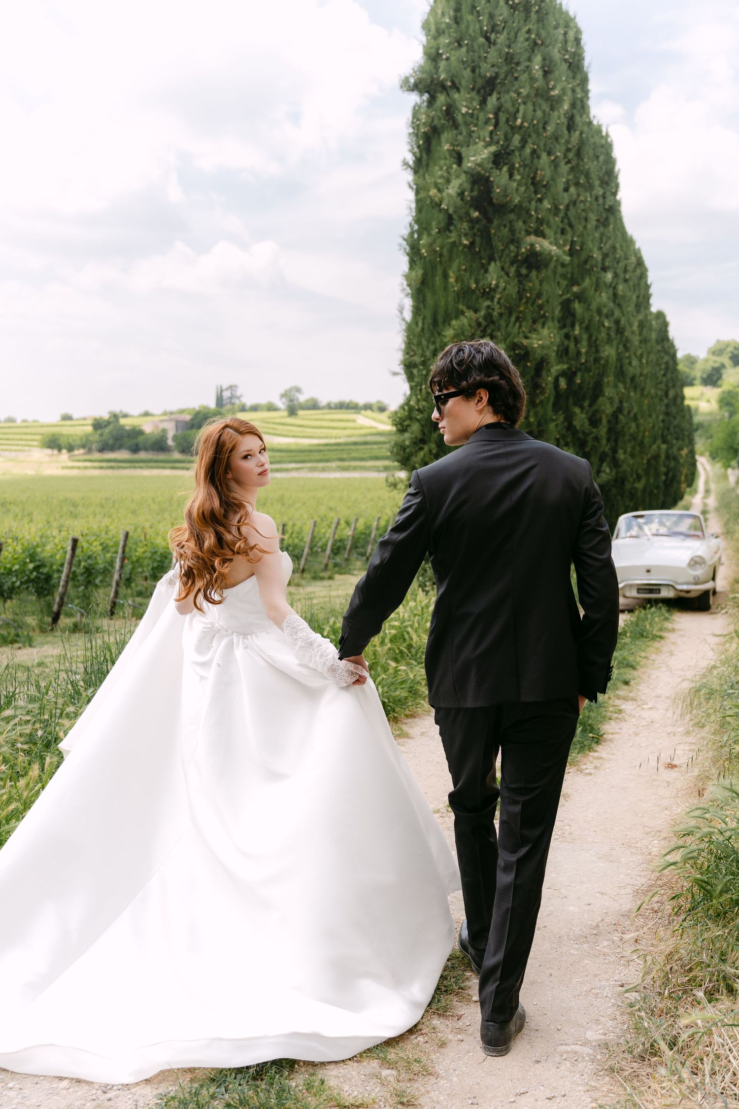 A couple in wedding attire walking along a countryside path lined with cypress trees.