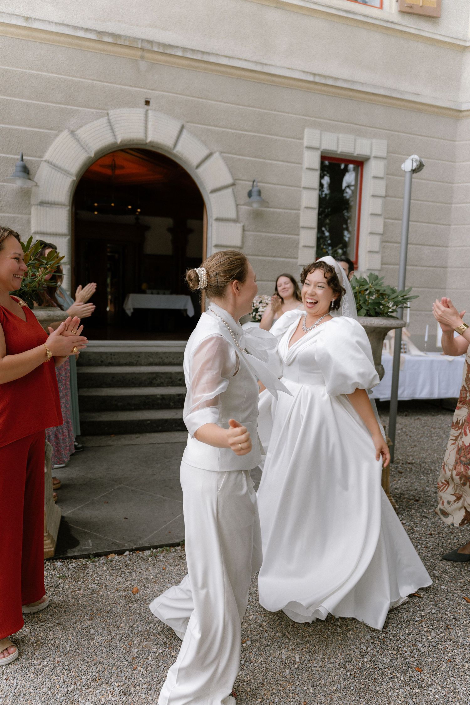 Two brides in white dancing joyfully outside a venue while guests in red applaud at a wedding celebration.
