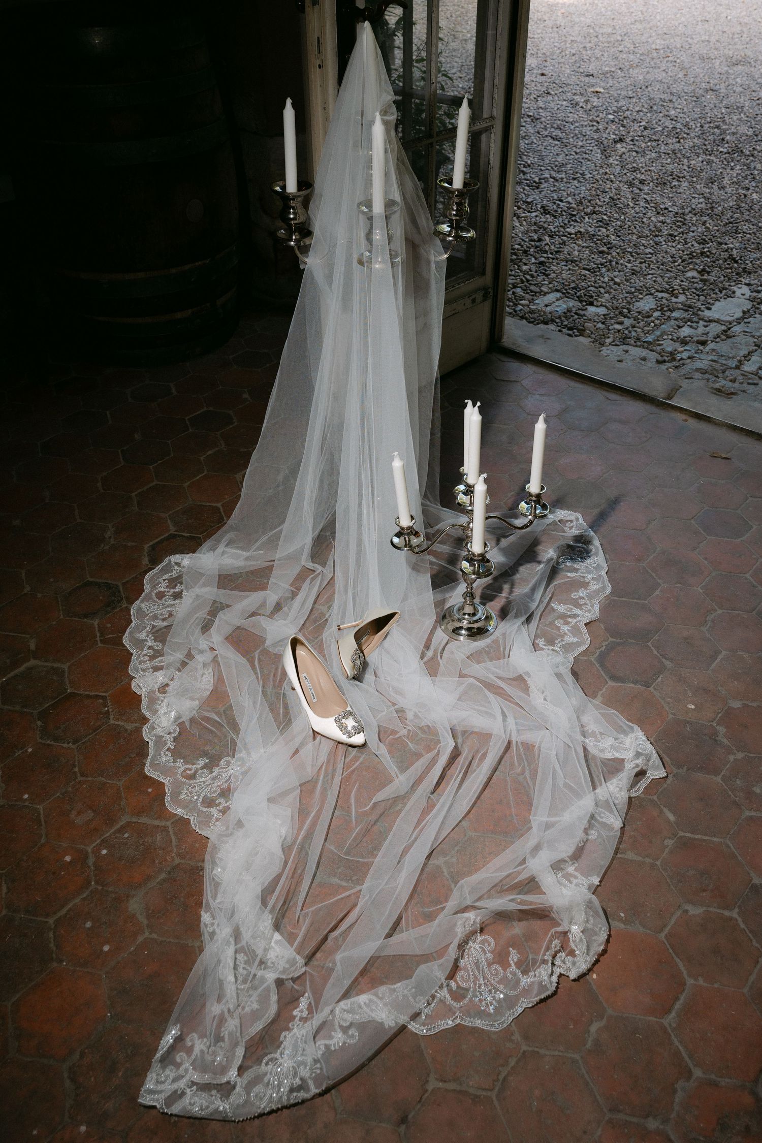 Long lace-trimmed bridal veil spread on a terracotta tile floor with white heels and candelabras holding lit candles near an open doorway.