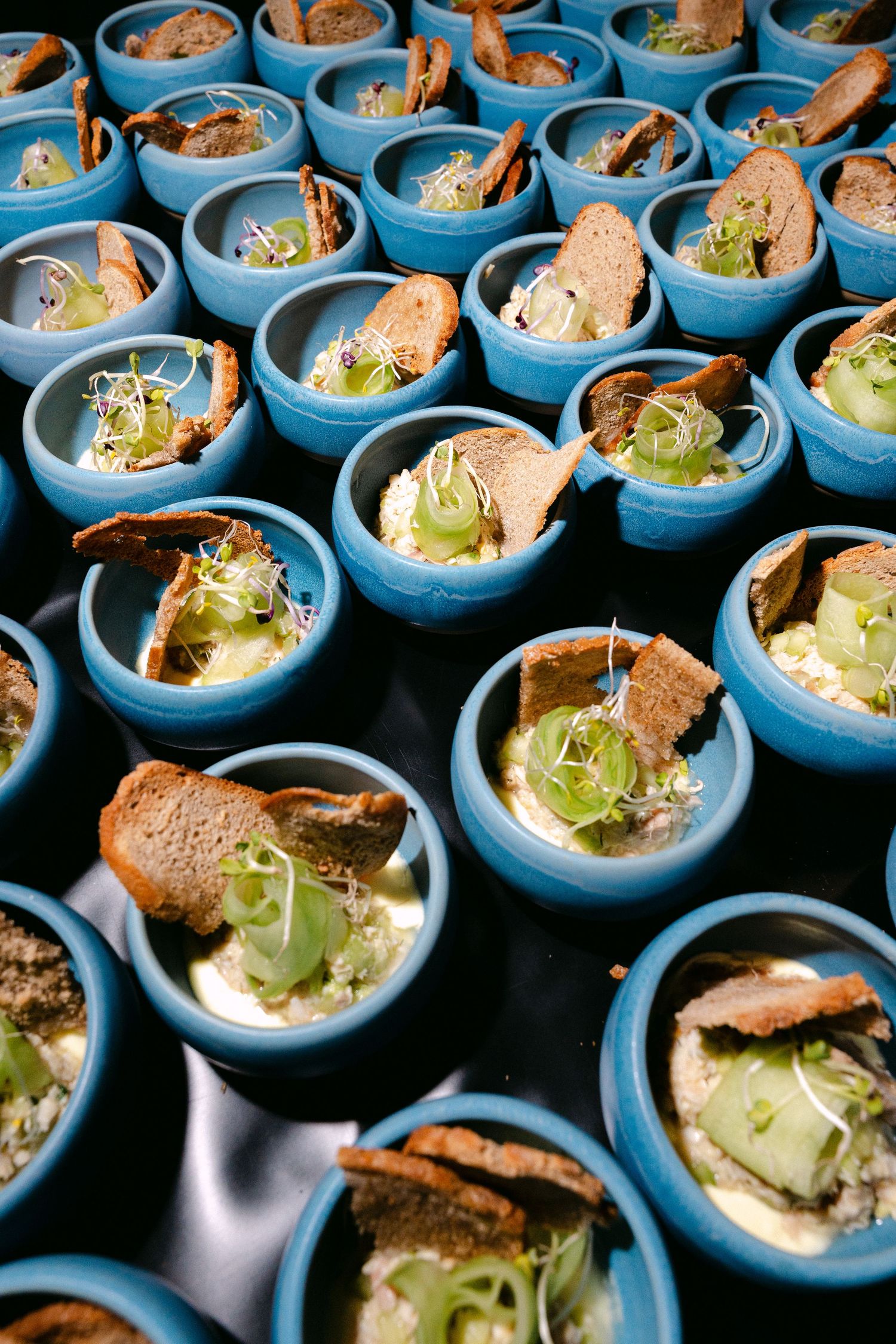 Rows of blue ceramic cups filled with elegant appetizers featuring bread, avocado, and microgreens at a catered event.