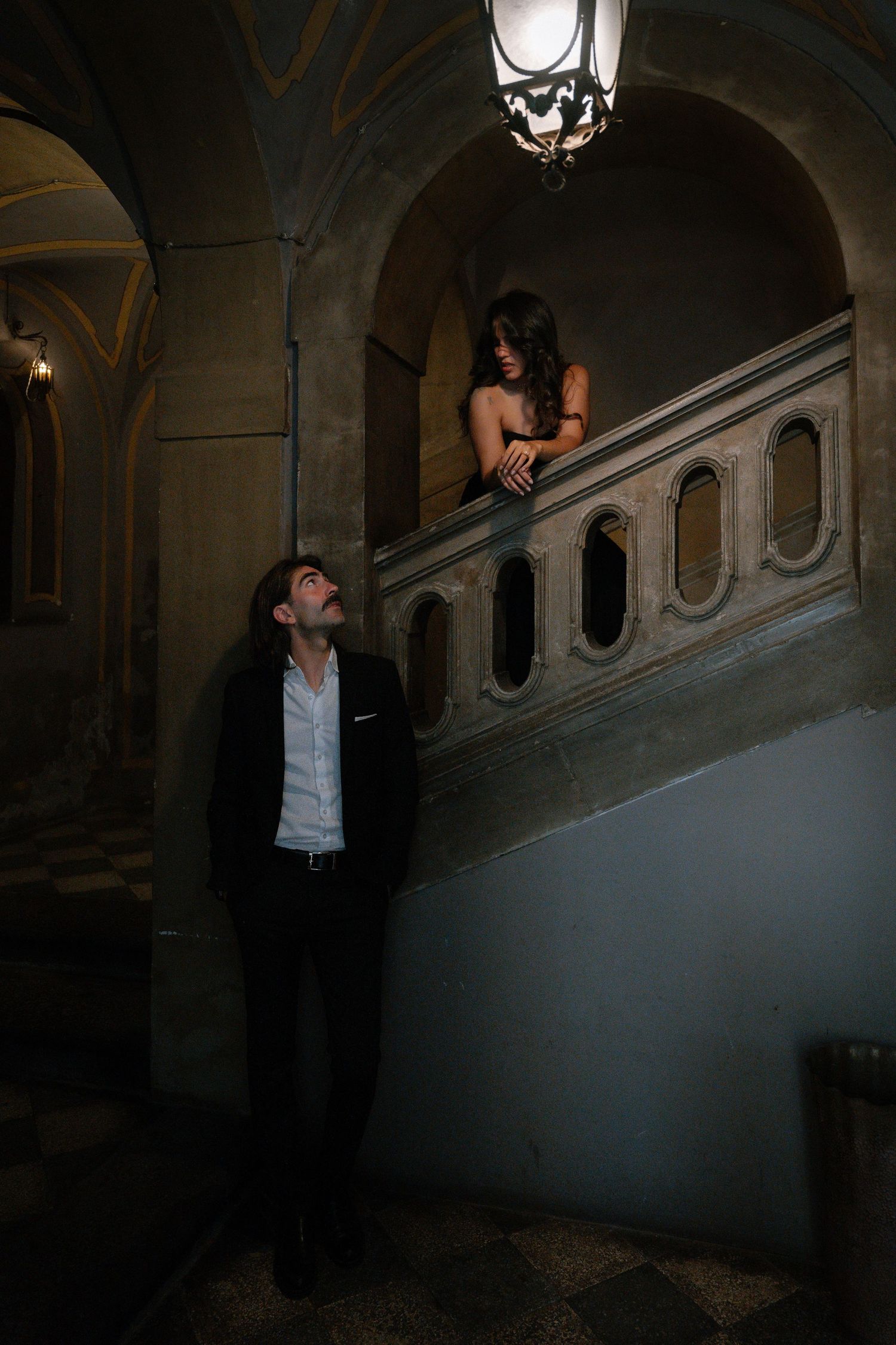 A man in a suit gazes up at a woman on an ornate staircase in a dimly lit historic building.