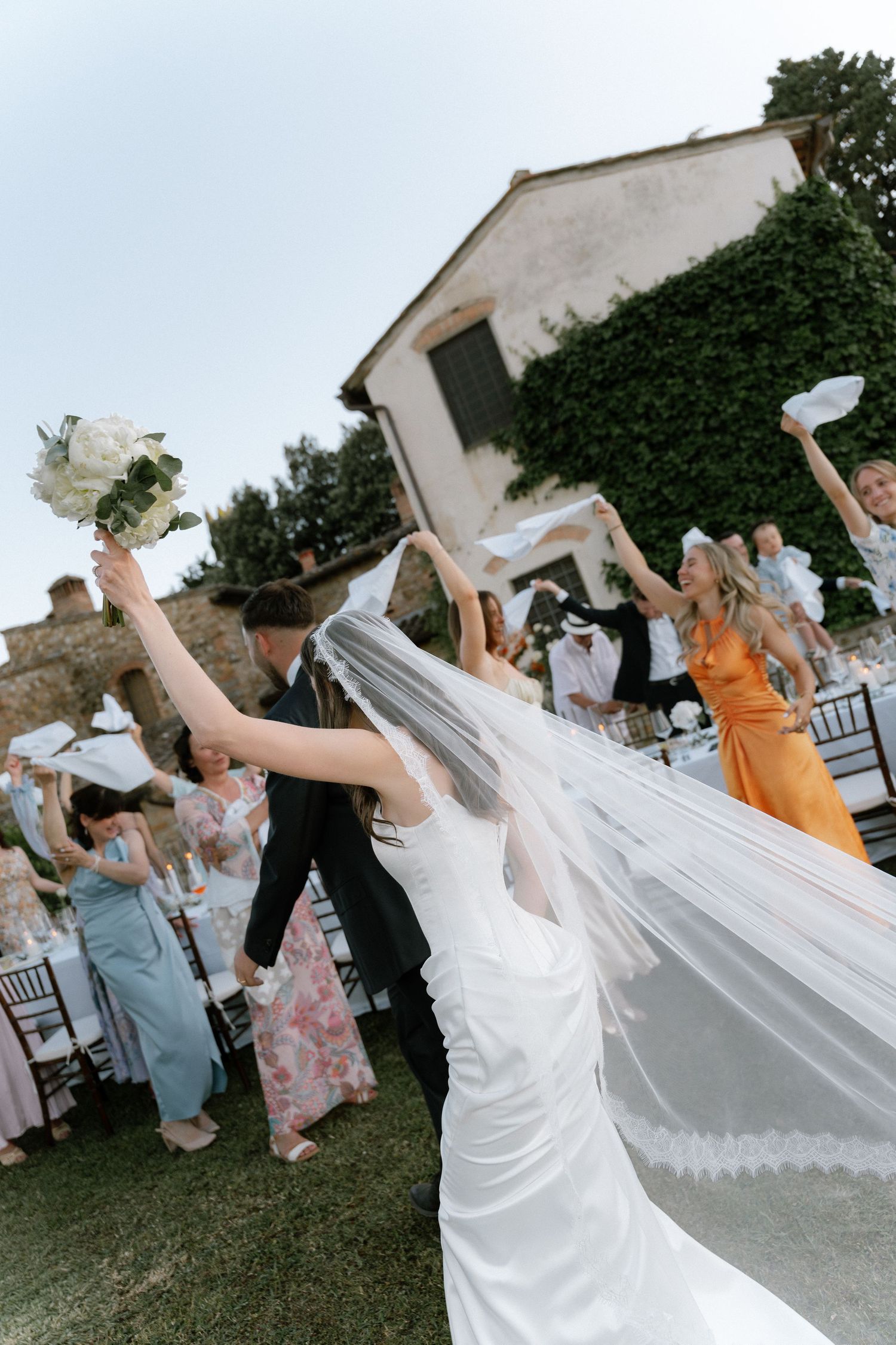 Bride in white gown and veil celebrates with guests outdoors at a wedding reception, raising bouquet joyfully.