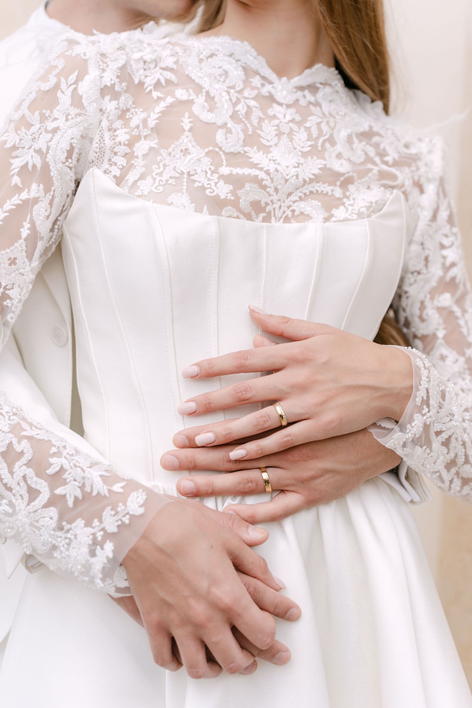 Close up detail of white lace wedding dress with long sleeves and delicate floral embroidery, showing hands with rings. Villa Mosconi Bertan