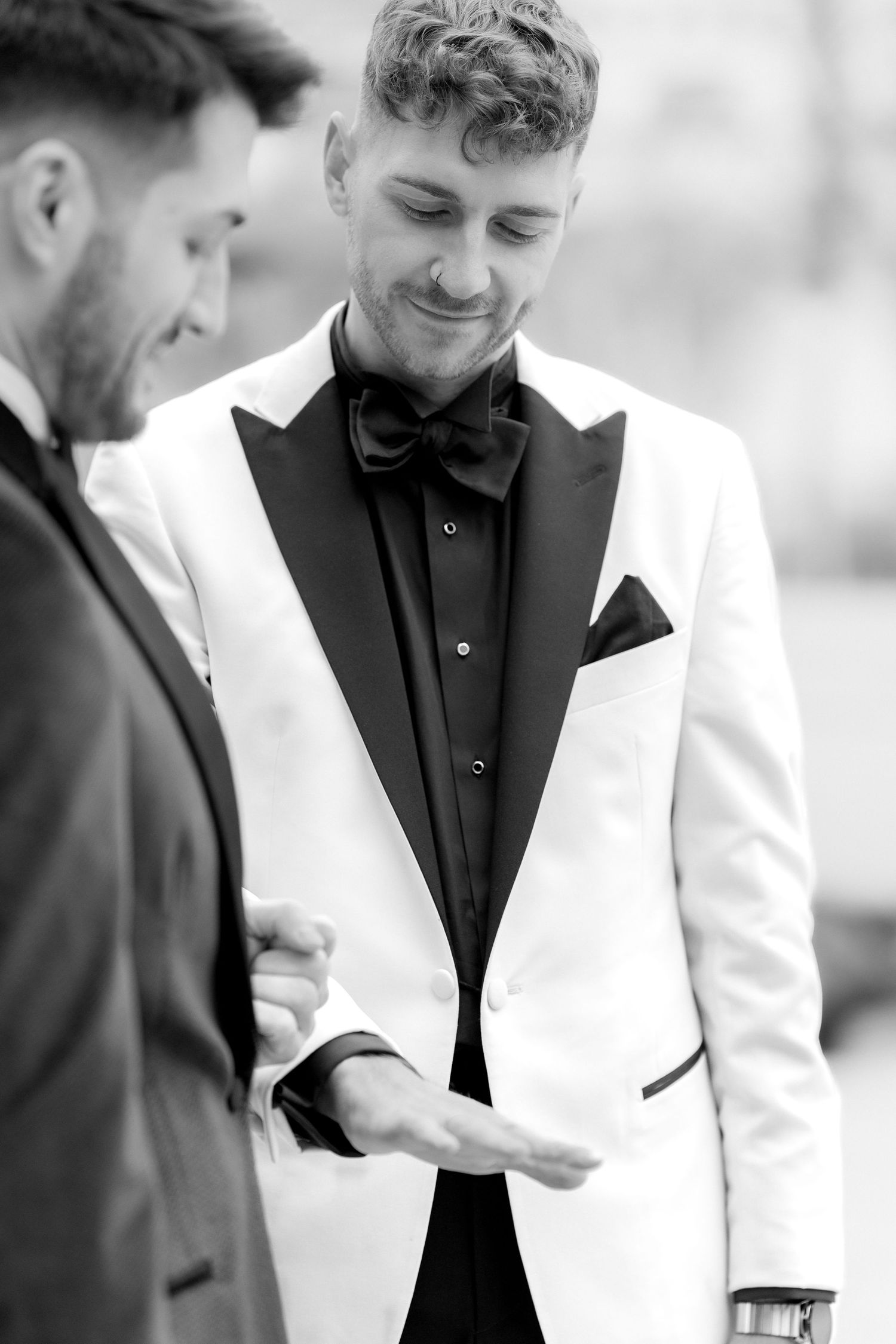 A groom in a white dinner jacket with black lapels and bow tie smiles while looking down at wedding preparations.