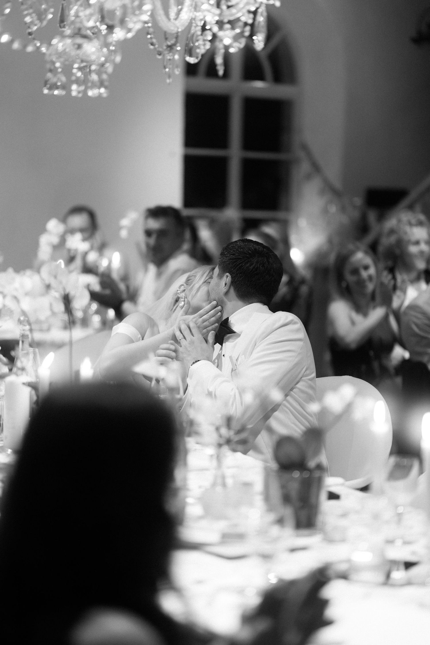 A black and white wedding reception scene with an elegant chandelier and guests seated at decorated tables.