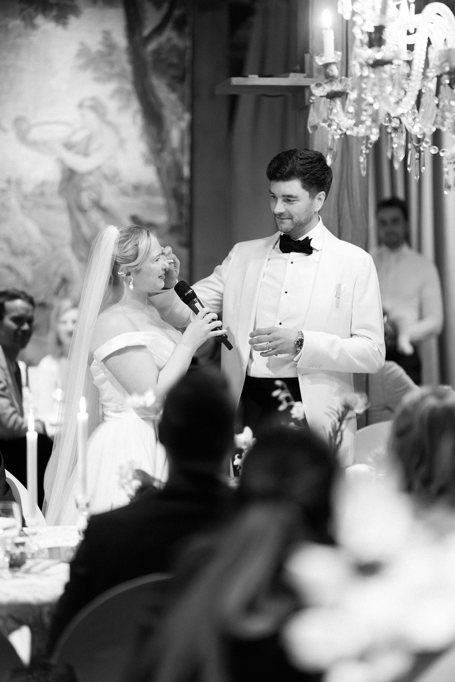 A black and white photo of a bride and groom sharing a toast at their wedding reception under an elegant chandelier.