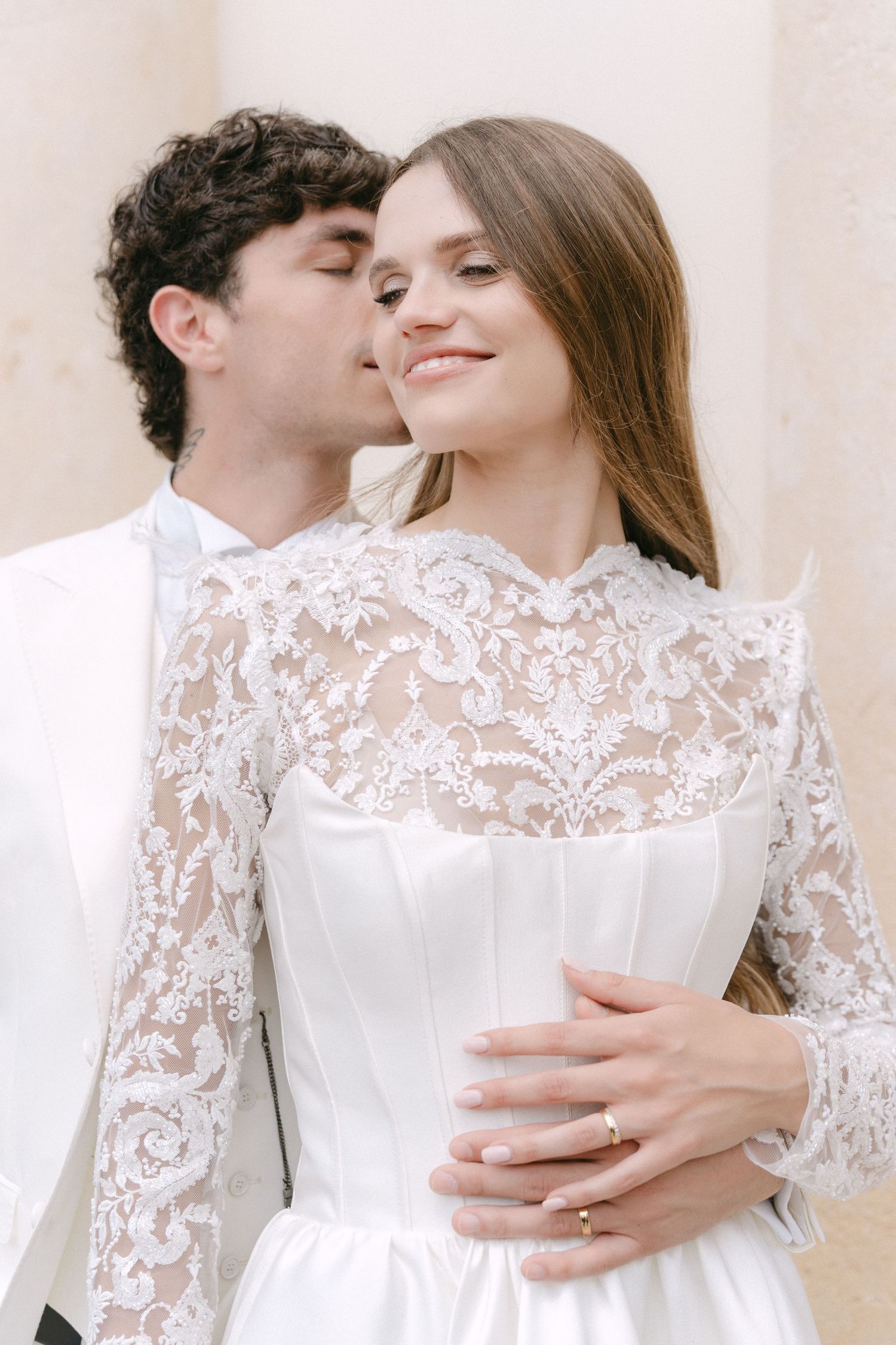 A romantic couple embraces in white lace wedding attire against a light background. Villa Mosconi Bertani