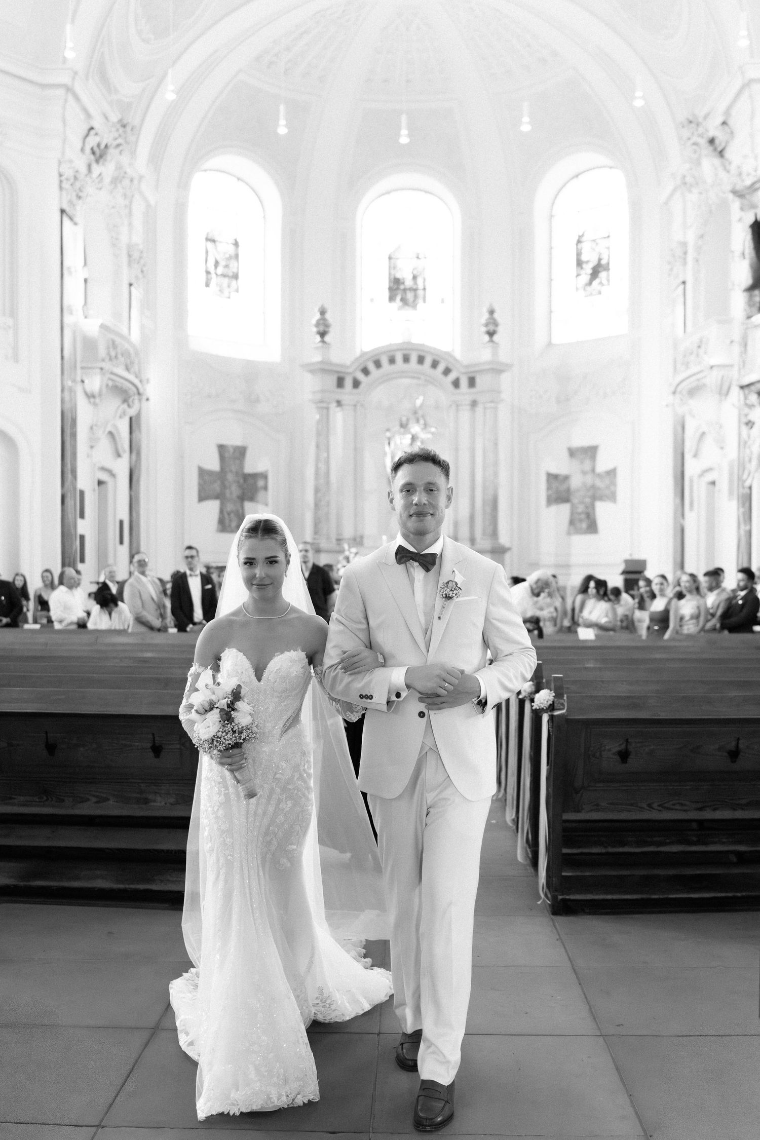 A bride and groom walk down the aisle of an elegant white church interior during their wedding ceremony.