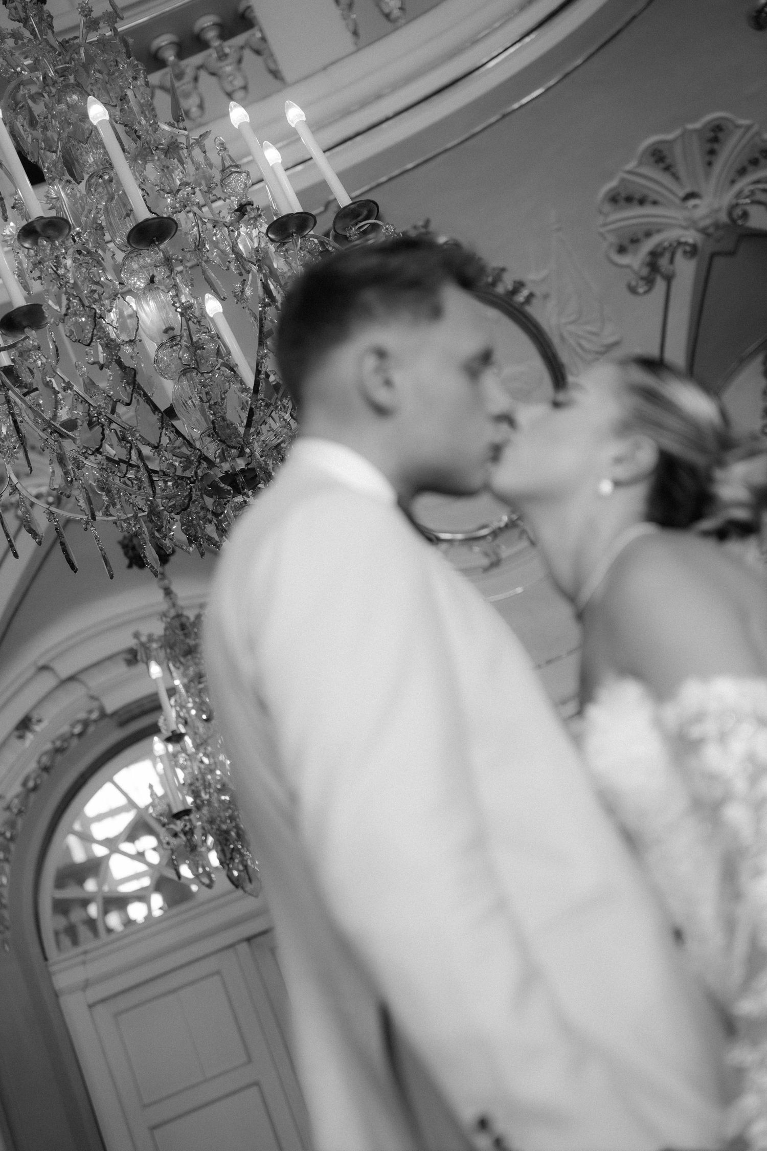 Black and white wedding photograph showing a romantic kiss under an ornate chandelier in an elegant venue. Schloss Ludwigsburg