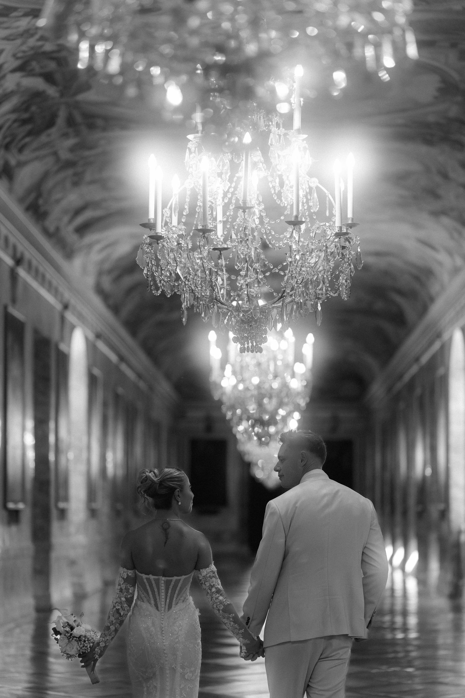 A romantic couple stands beneath ornate crystal chandeliers in an elegant palace hallway. Schloss Ludwigsburg