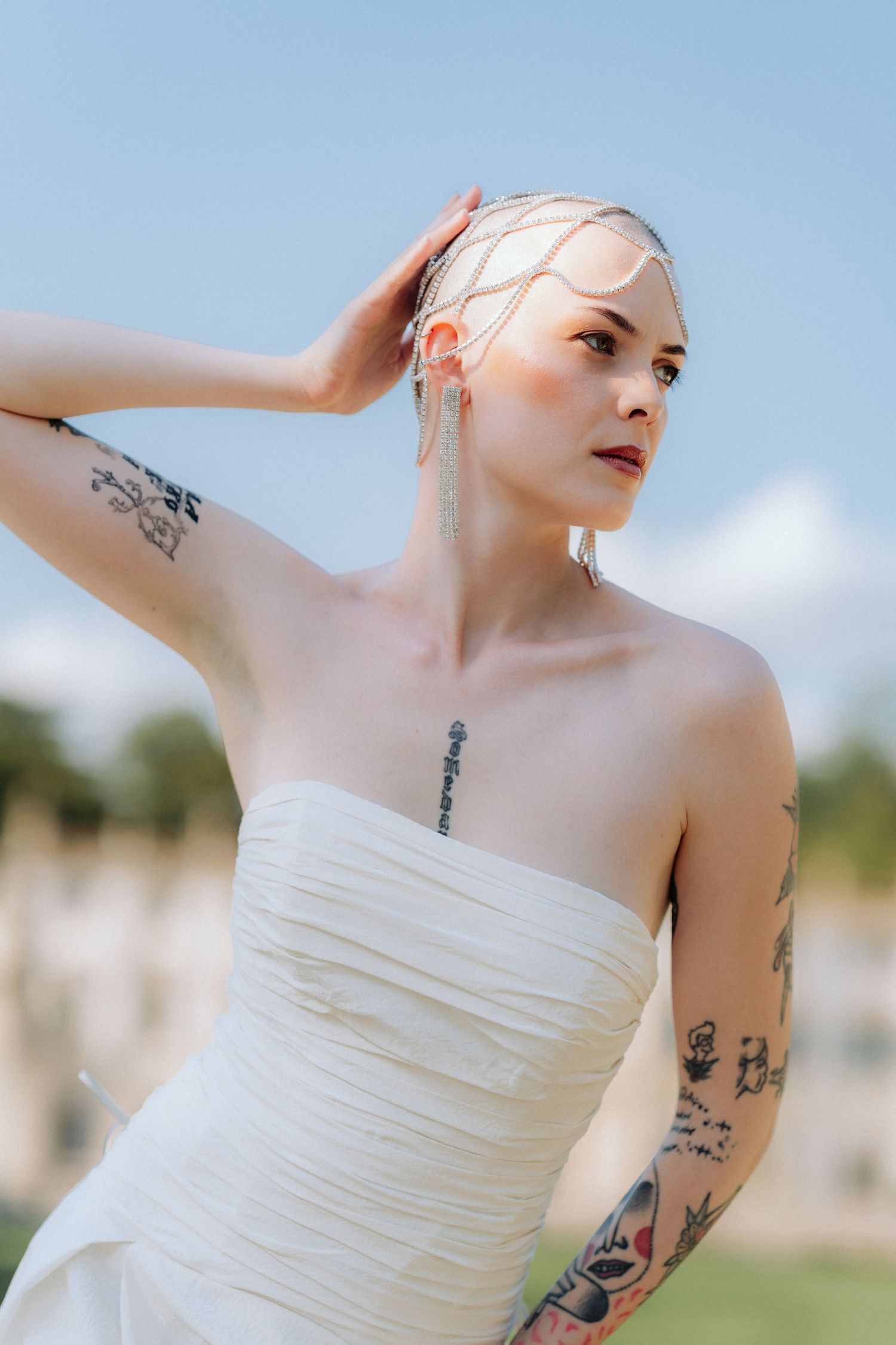 Person in white strapless wedding dress poses outdoors with visible arm tattoos against blue sky background.