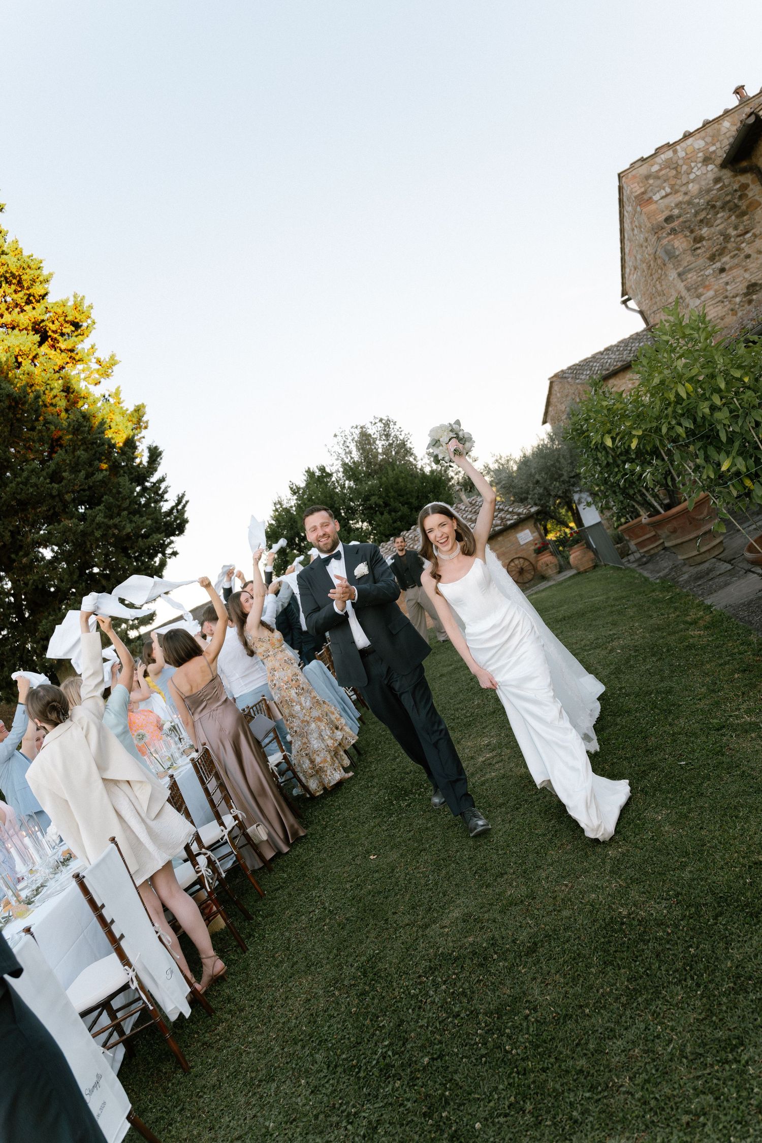 Bride and groom walk joyfully down outdoor aisle at Tuscan villa wedding, guests cheering with raised glasses.