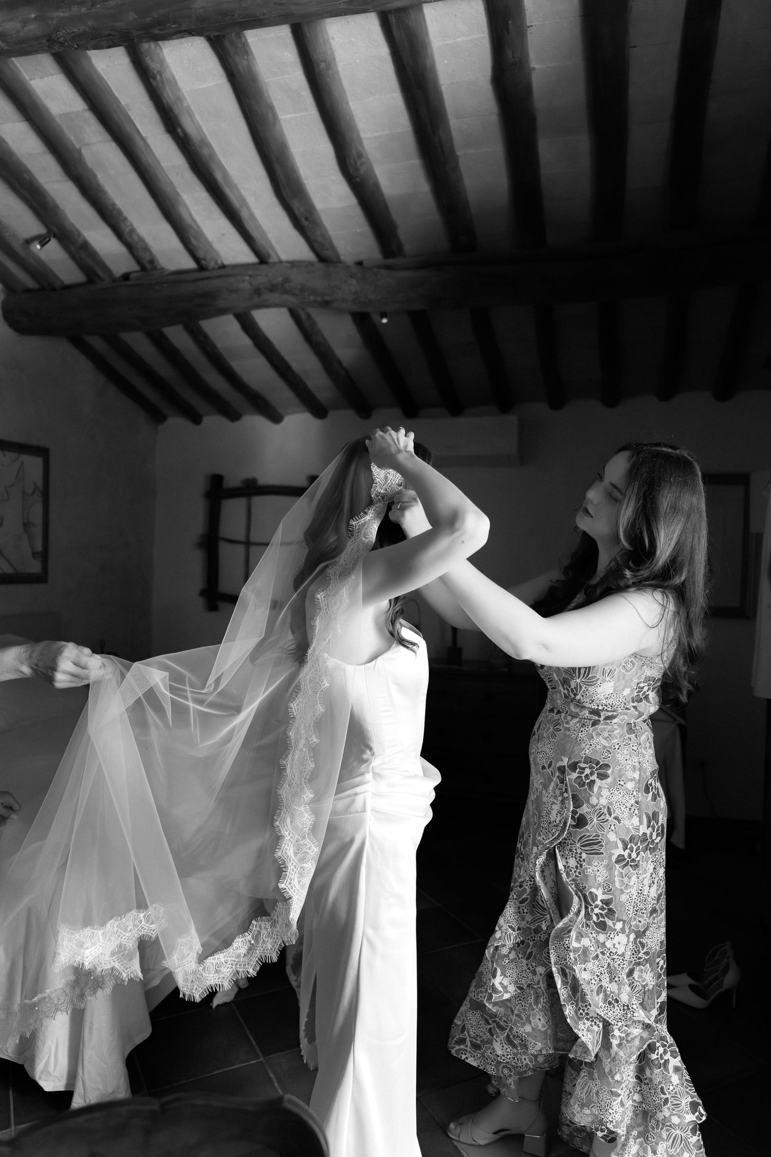 Black and white photo of a bride getting her veil adjusted by a bridesmaid in a rustic venue.