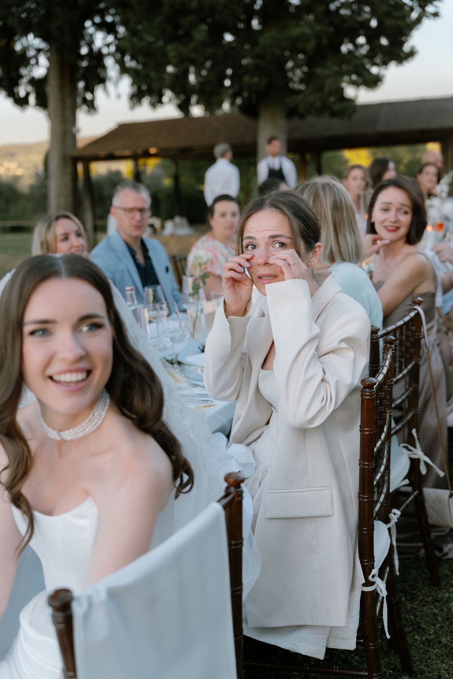 Emotional wedding guest in white blazer wiping tears at outdoor reception dinner table.
