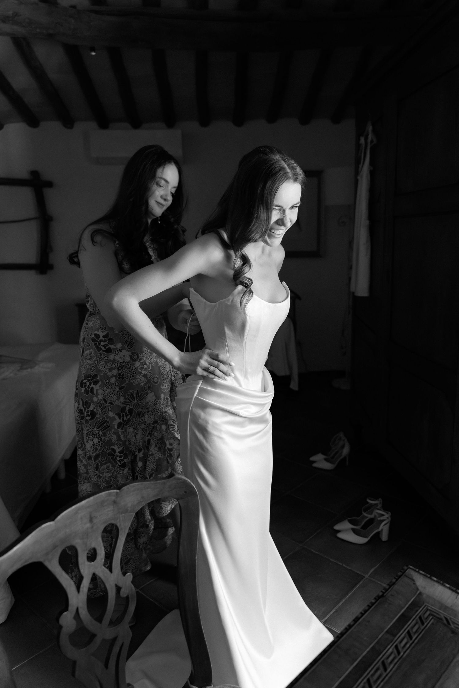 Black and white photo of a bride in a sleek white gown being helped by a bridesmaid in a rustic room.