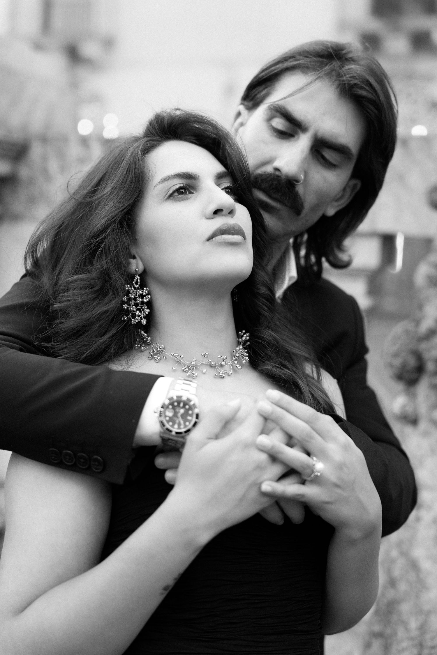 Black and white photo of an elegant couple embracing, woman in black dress with diamond necklace, man in suit with watch.