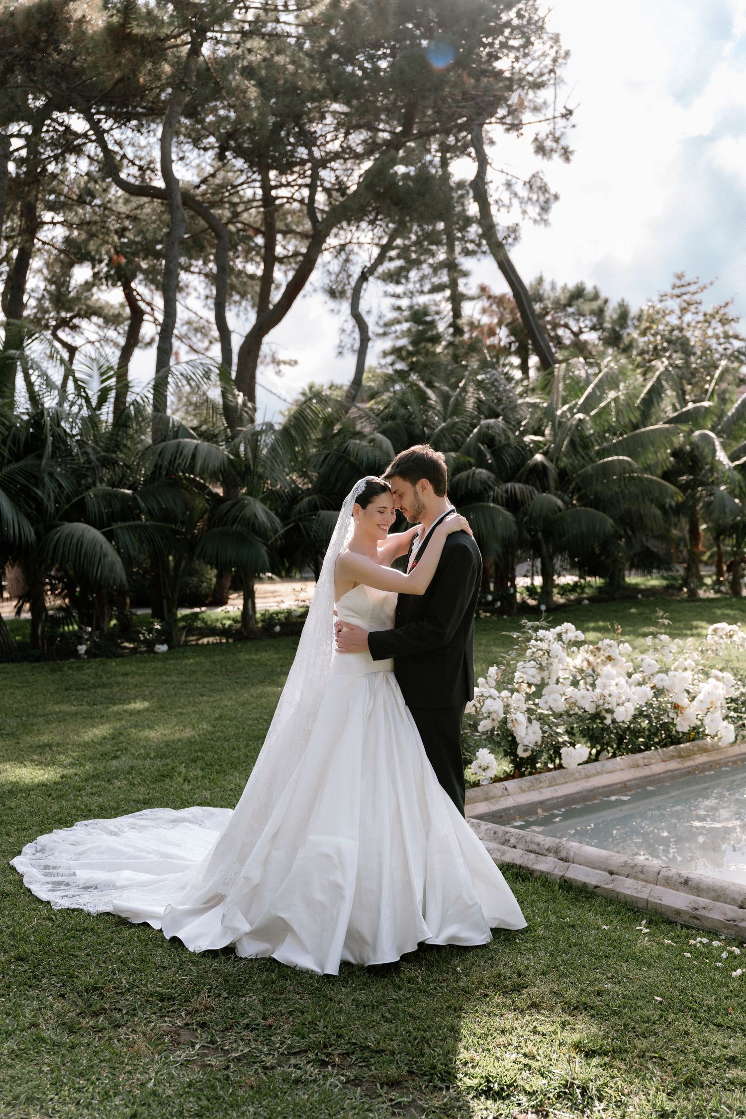 Bride and groom embrace in lush garden, white wedding gown with long train, groom in black suit, surrounded by trees.
