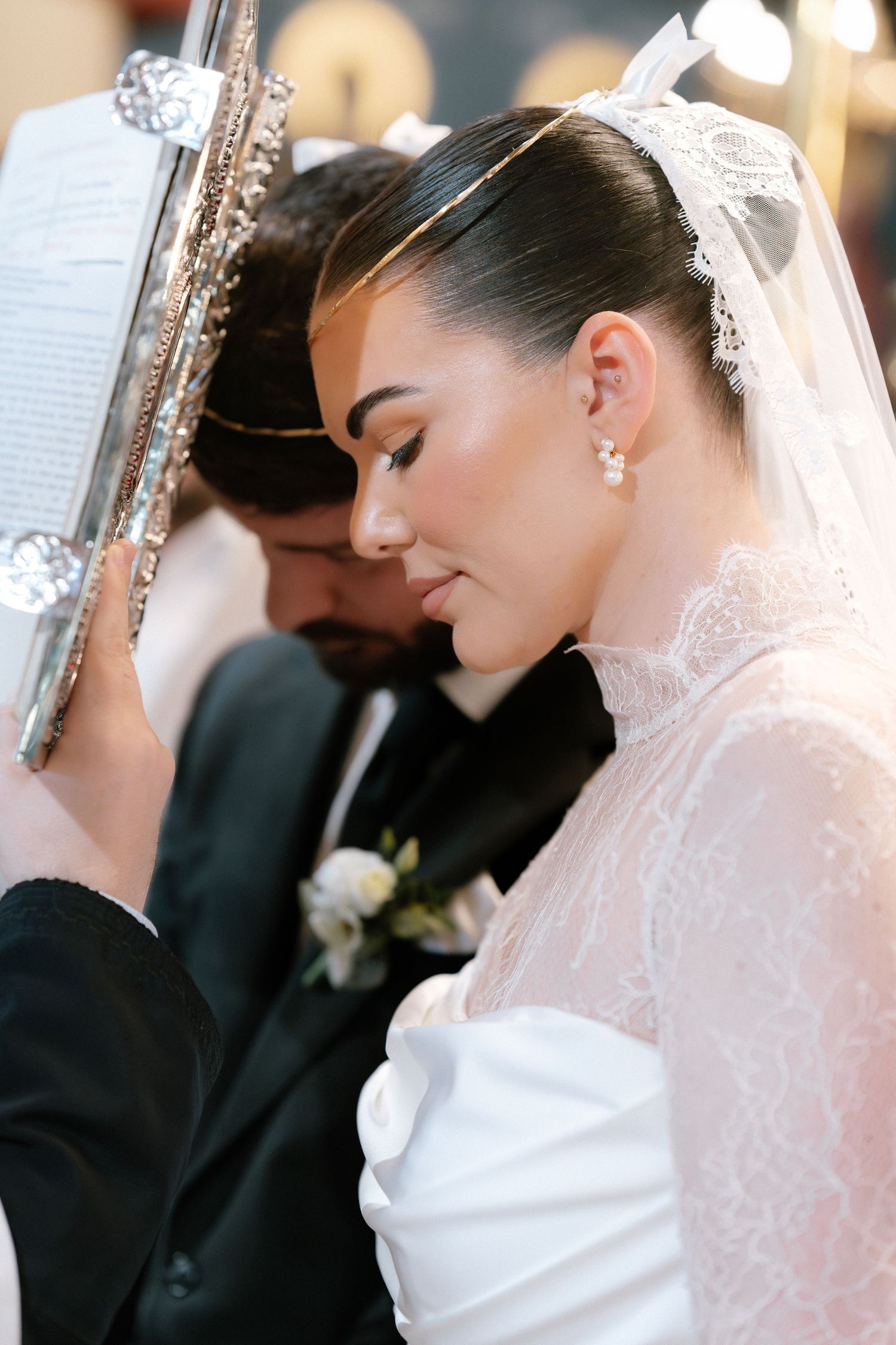 Bride in lace veil and groom in black suit share a solemn moment during a wedding ceremony.