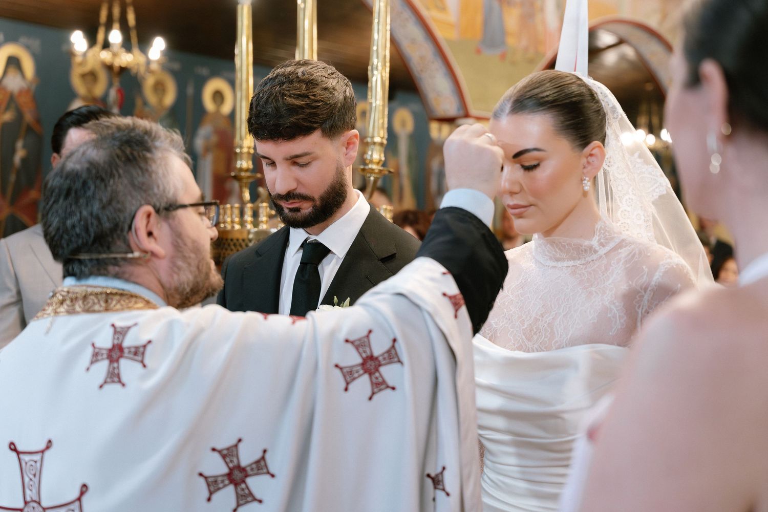 Orthodox wedding ceremony with priest blessing couple in white lace gown and black suit inside decorated church.