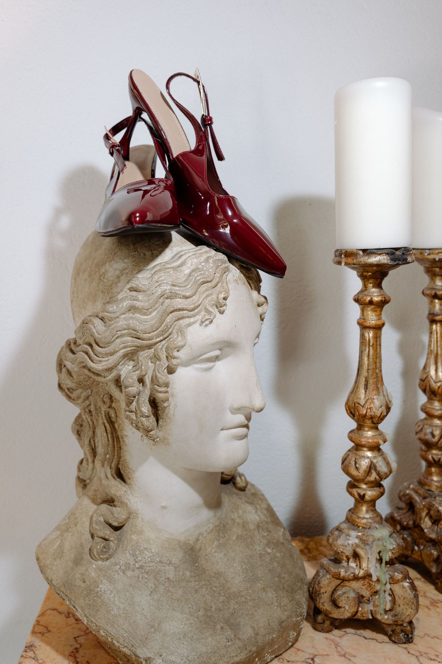 Dark red patent leather slingback heels displayed on a classical marble bust sculpture near golden candlesticks.