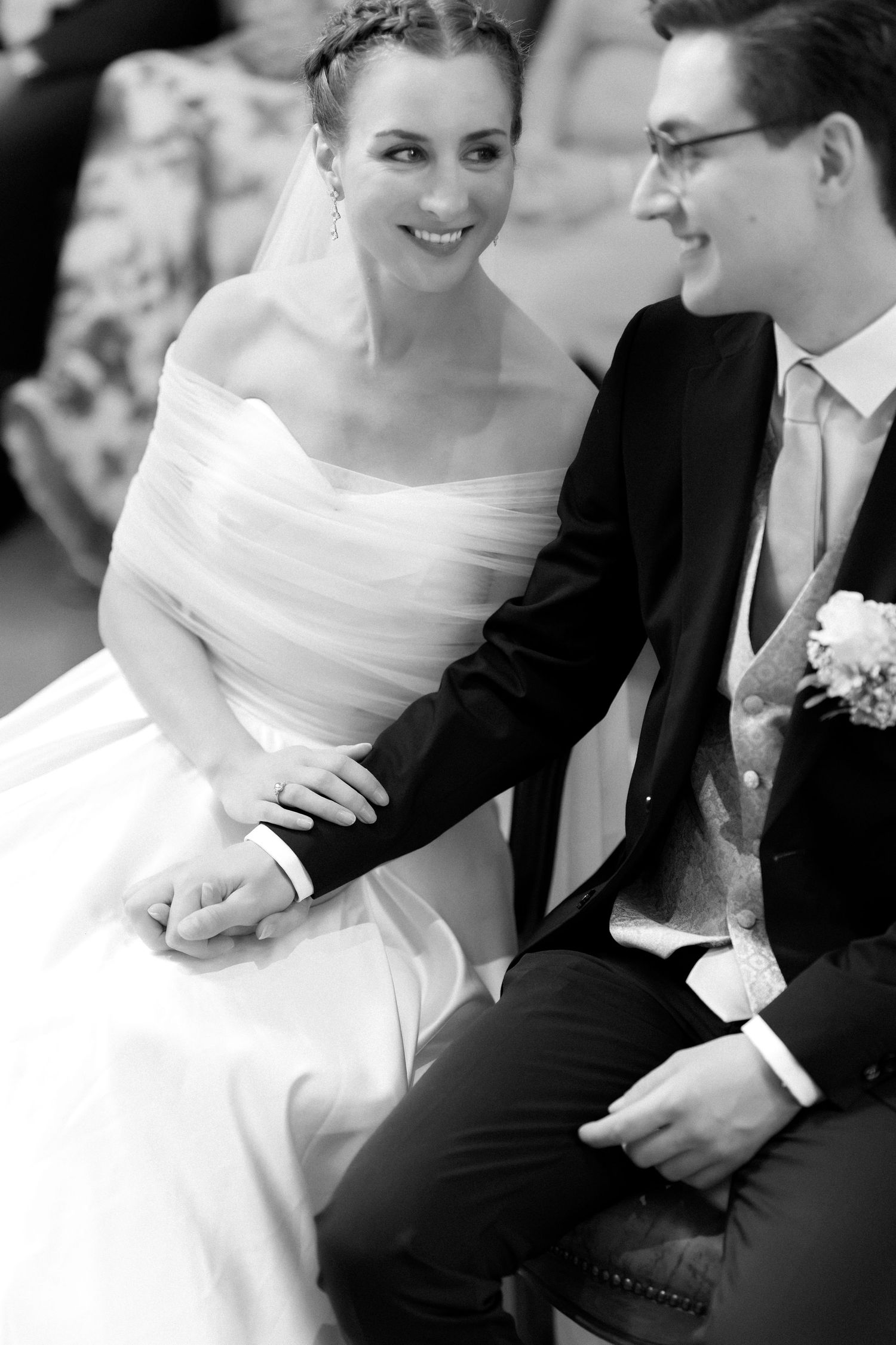 Smiling bride in an off-the-shoulder wedding gown and veil holds hands with a groom in a suit and boutonniere during their ceremony.