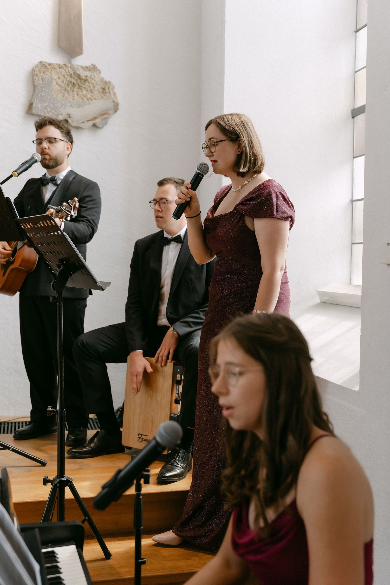 Wedding band performing live in a bright chapel, with a singer in a burgundy dress, a guitarist, a cajón player and a pianist.