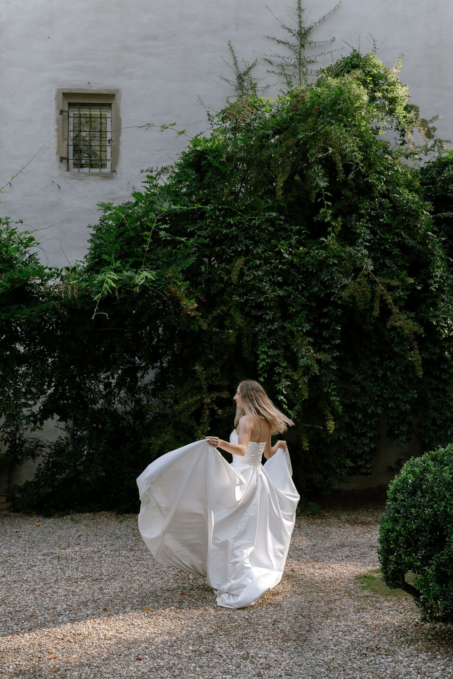 Bride in a flowing white wedding gown twirls on a gravel courtyard beside lush green ivy and an old stone building.