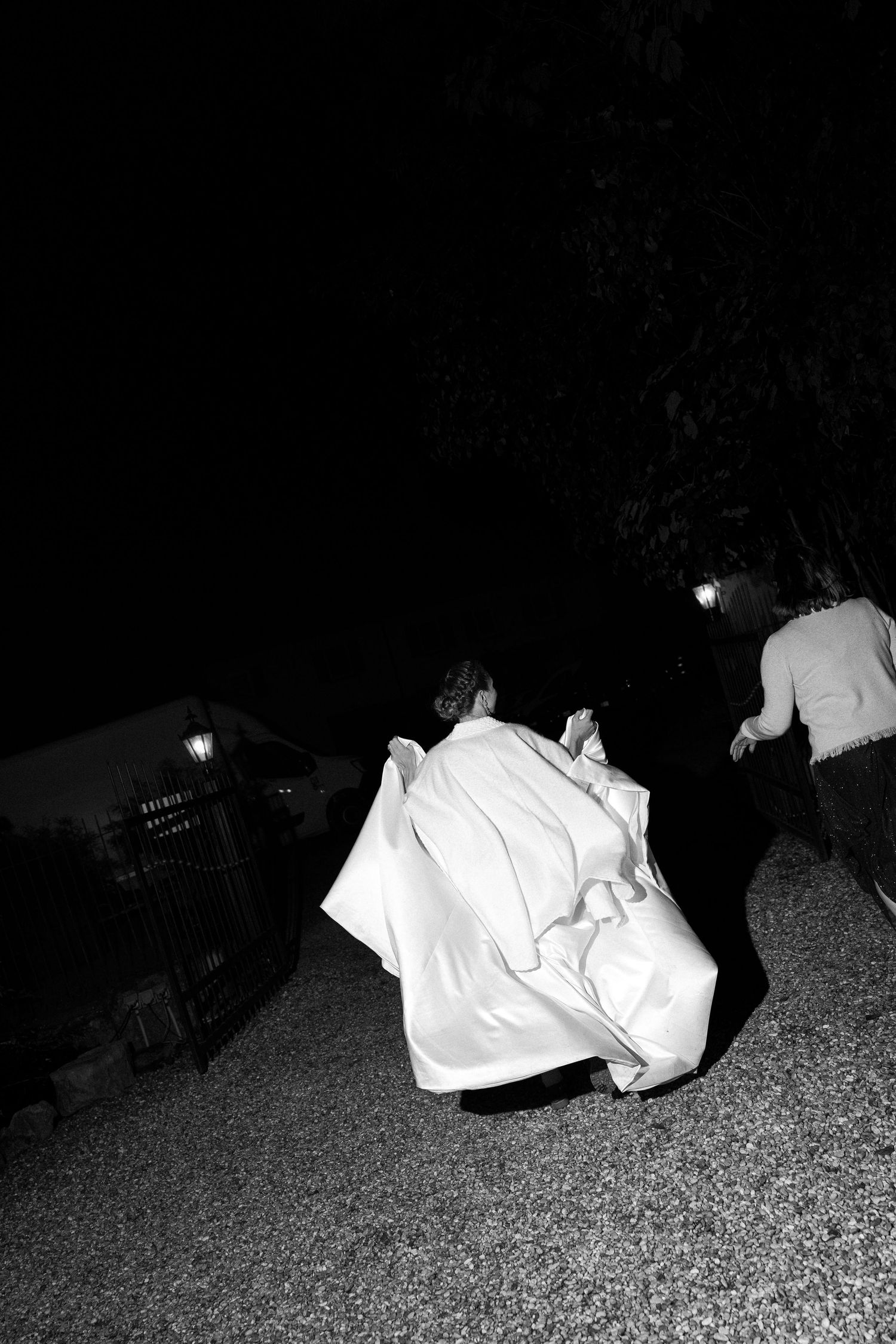 Black and white photo of a bride in a flowing white gown running along a gravel path at night as a guest follows nearby.