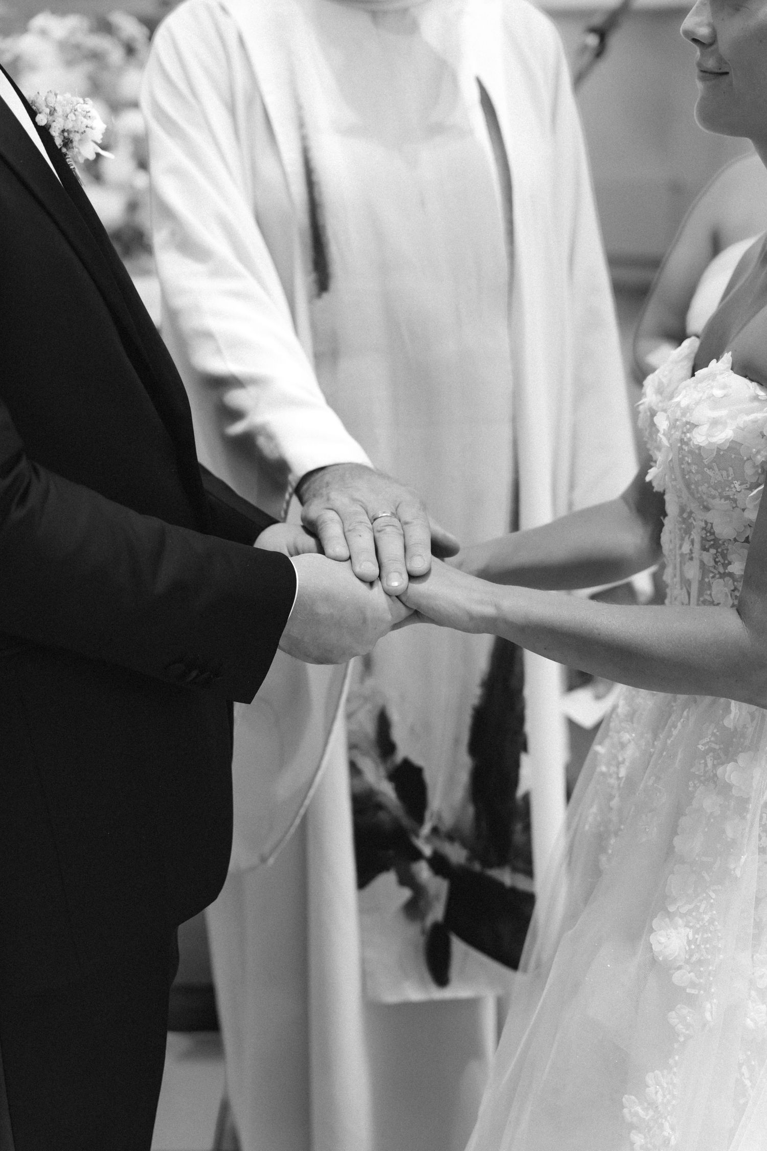Black and white photo of a bride and groom joining hands as an officiant in a robe blesses their wedding ceremony.