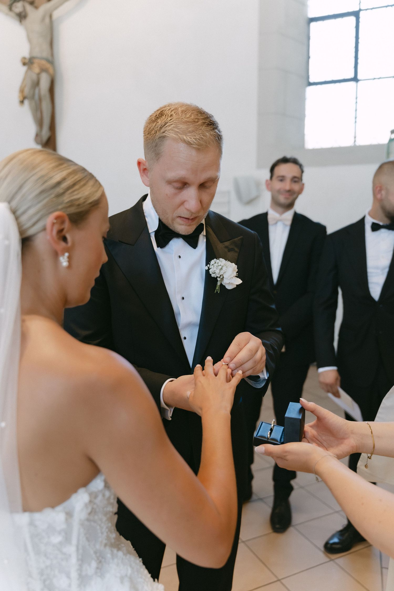Groom in a black tuxedo and bow tie places a ring on the bride's finger during a church wedding ceremony as groomsmen look on.