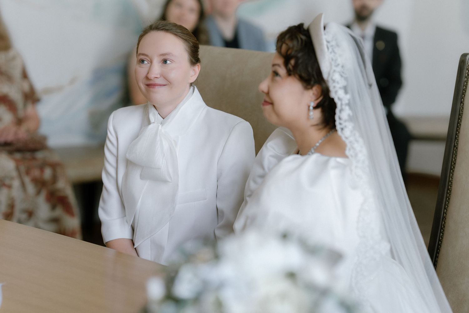 Two brides in white wedding attire smile joyfully during their ceremony, one in a suit and one in a traditional gown.