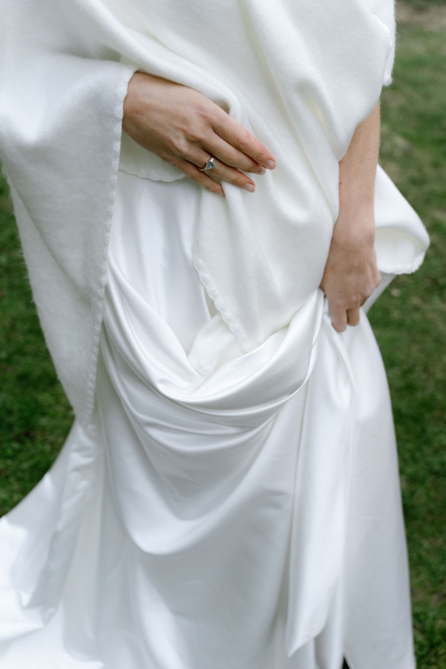 Bride in a flowing white satin gown and soft draped wrap with hands resting at her waist, a ring visible, standing on green grass.