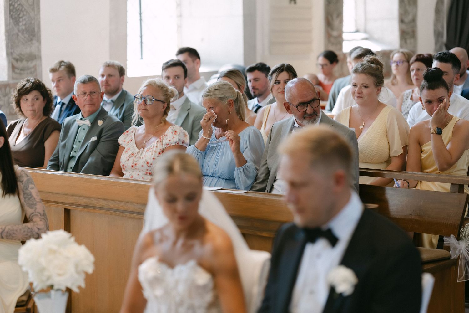 Wedding guests seated in church pews smile and tear up as a bride in a white lace gown and a groom in a tuxedo stand during the ceremony.