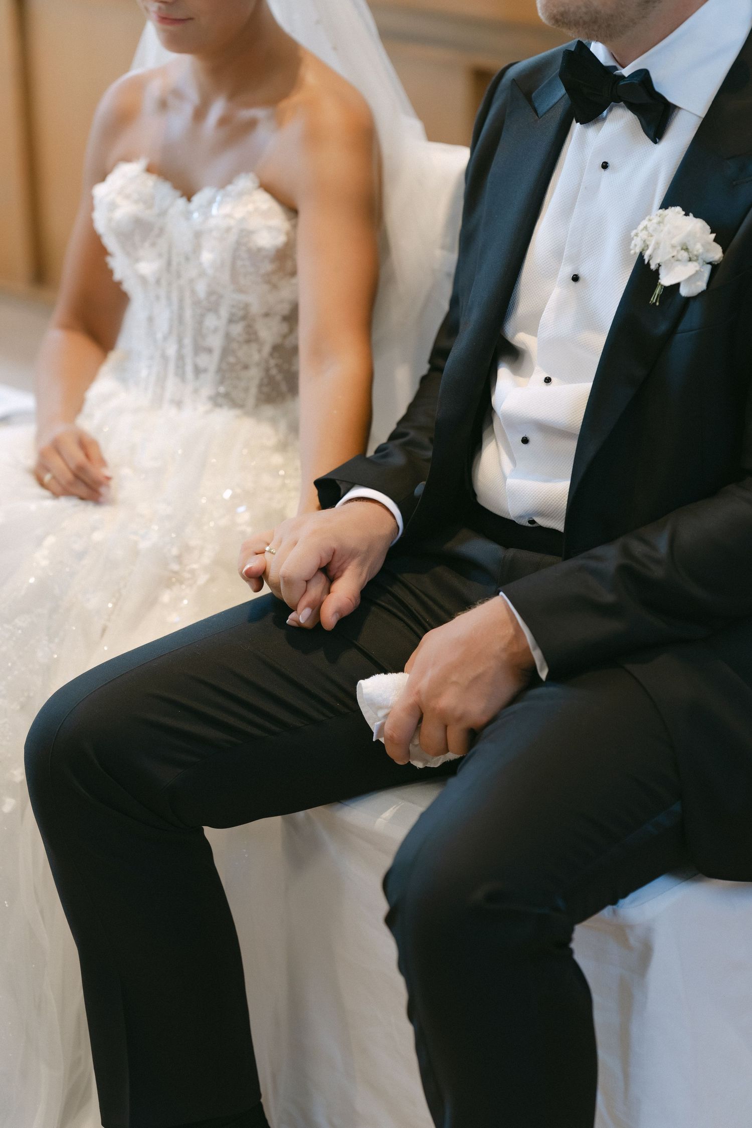 Bride in a strapless lace gown and veil sits holding hands with a groom in a black tuxedo and bow tie during a wedding ceremony.