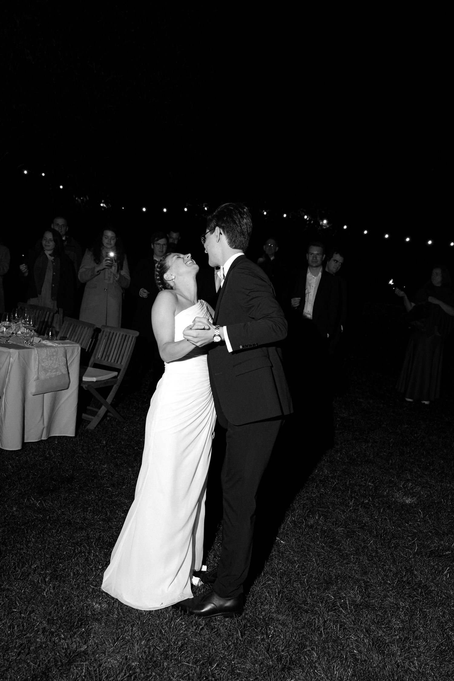 Bride in a white gown and groom in a dark suit share a first dance outdoors at night as wedding guests watch beneath string lights.