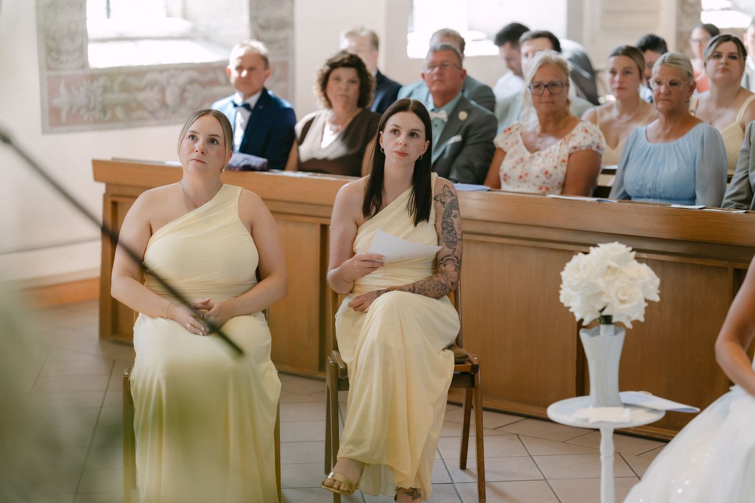 Two women in matching yellow satin dresses sit in a church pew while wedding guests look on, with white flowers in a vase nearby.
