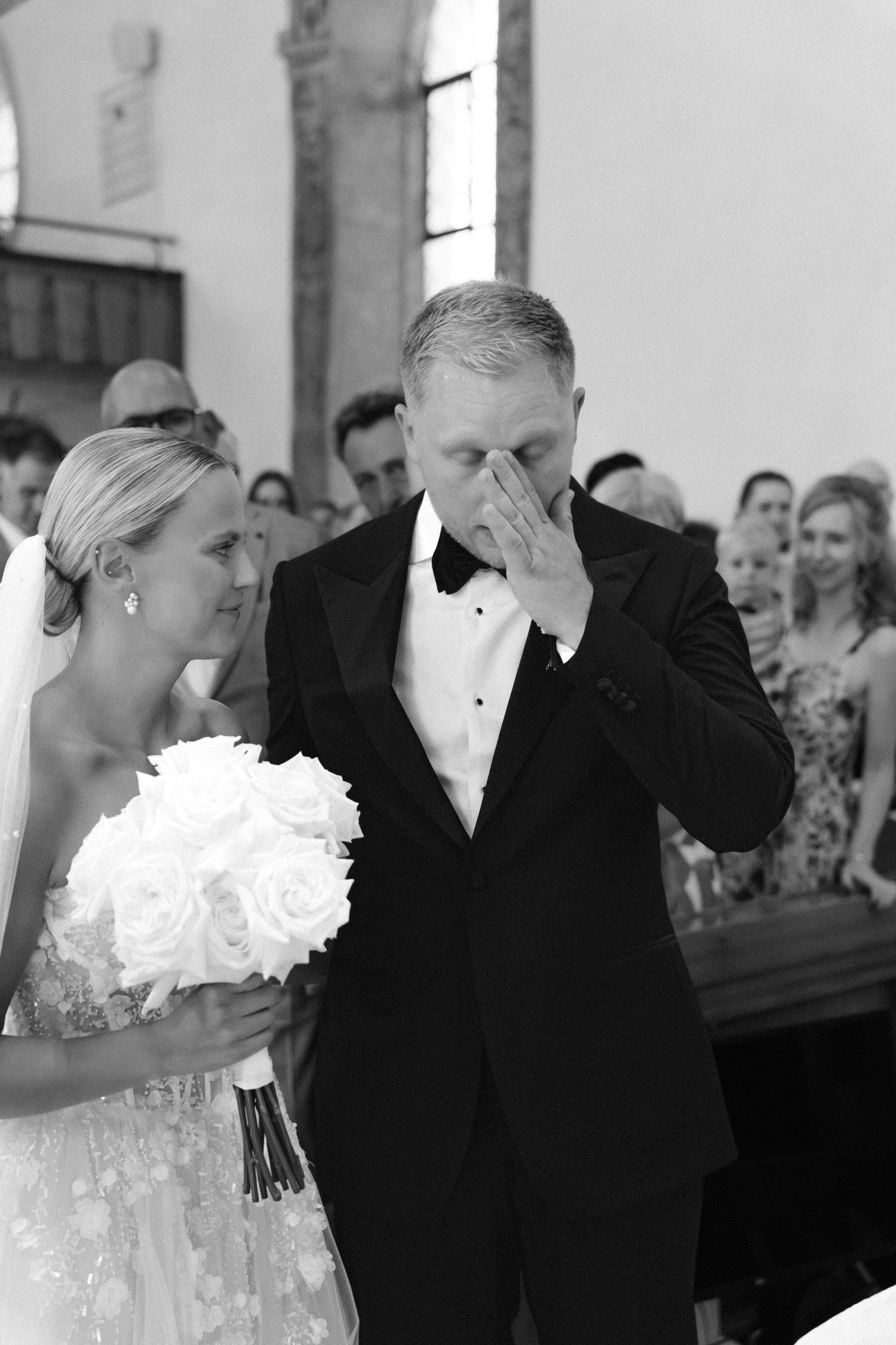 Emotional groom in a tuxedo and bow tie wipes his eyes beside a bride holding a white rose bouquet during a wedding ceremony.