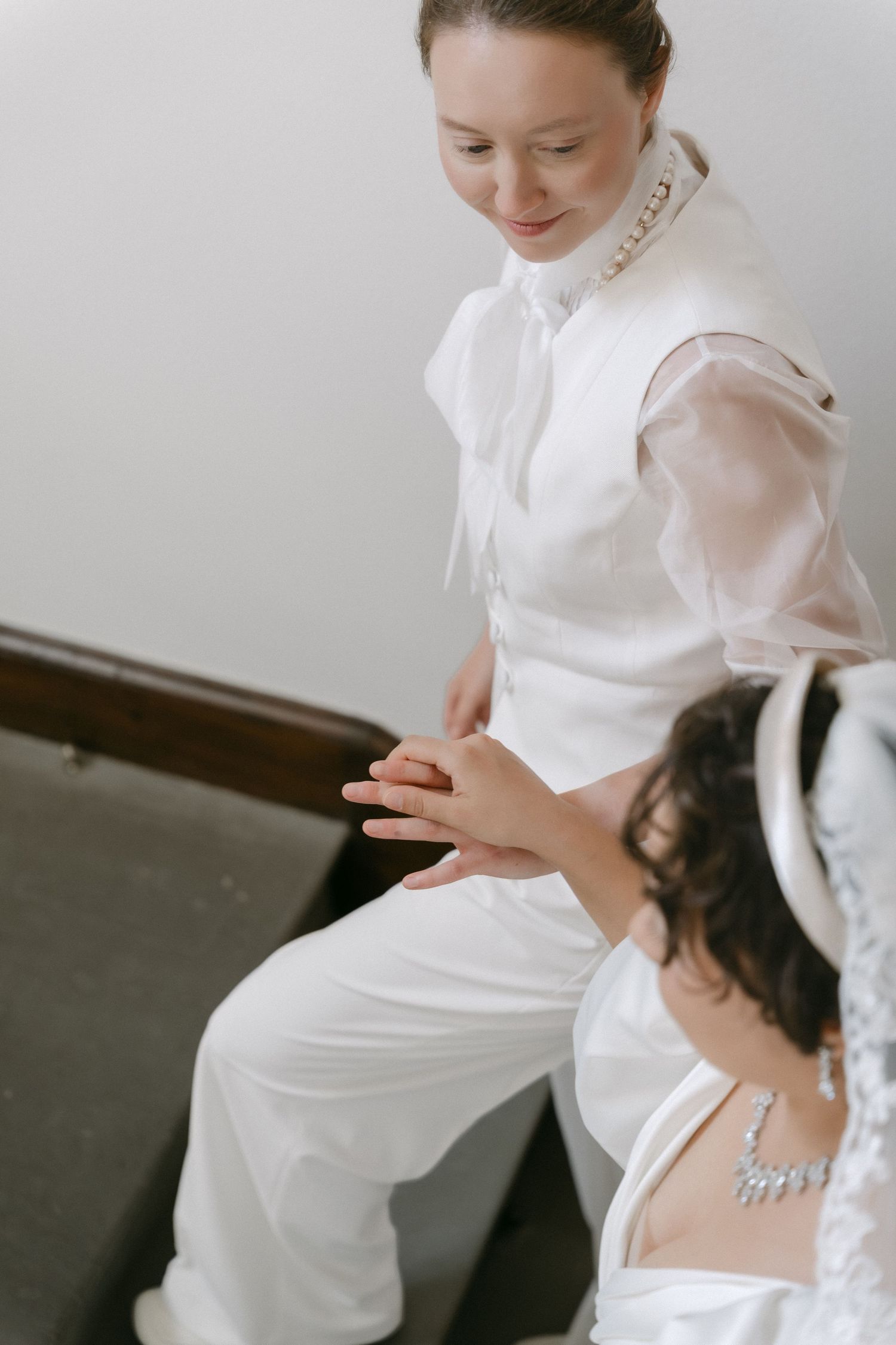 Two brides in white wedding attire hold hands on church steps, one wearing a pearl necklace and pantsuit.
