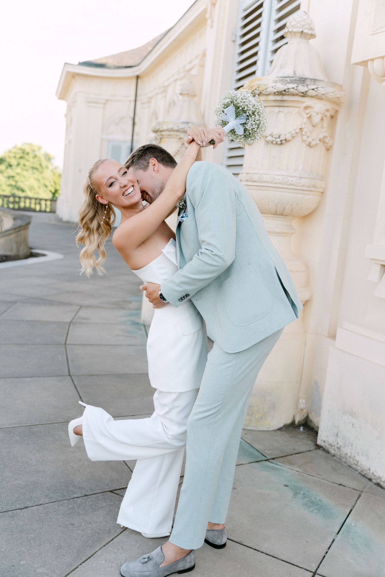 Romantic couple in light blue suit and white dress share intimate dance poses against ornate stone building exterior. Schloss Solitude