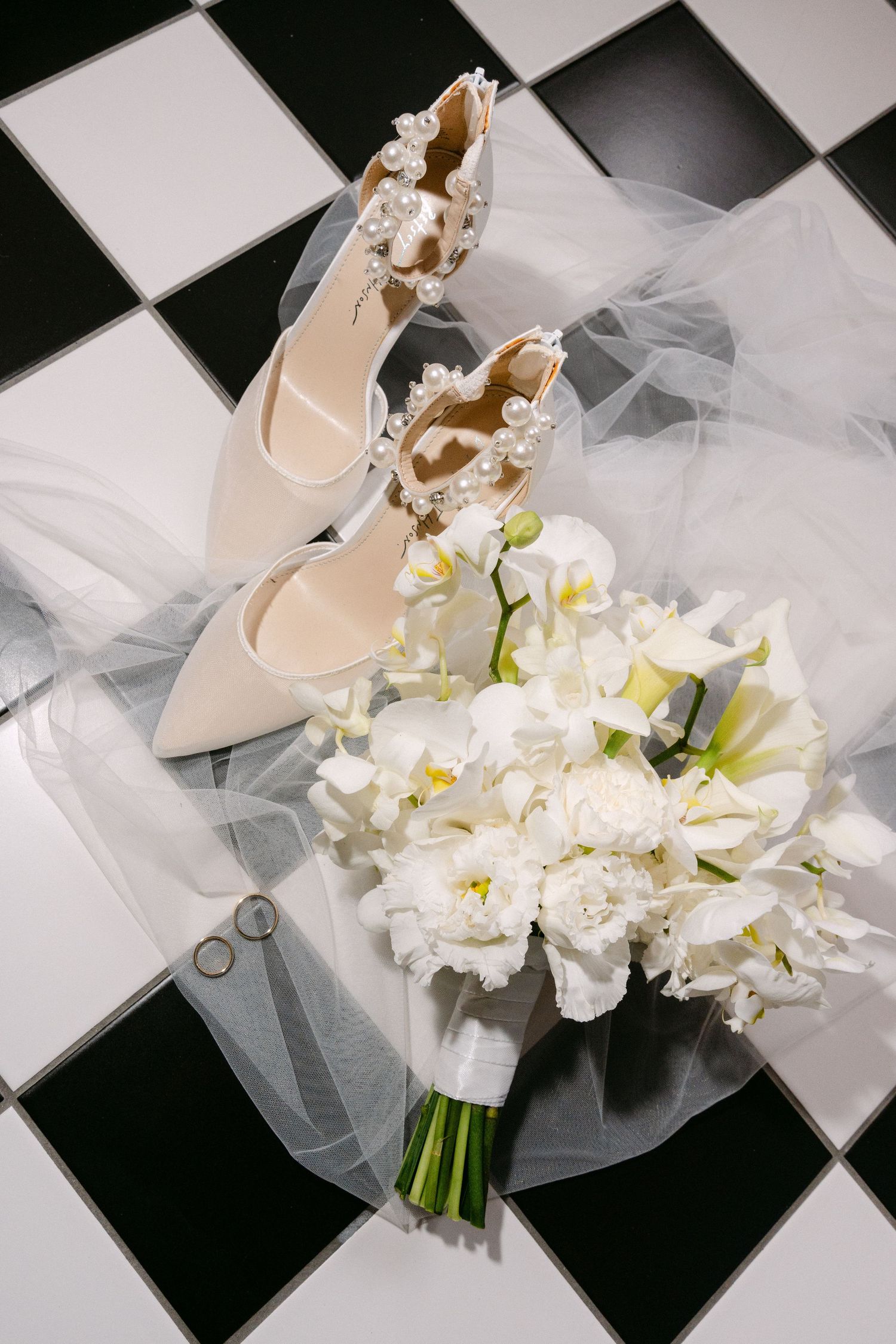Elegant bridal shoes and white floral bouquet arranged on black and white checkered surface with delicate tulle veil.