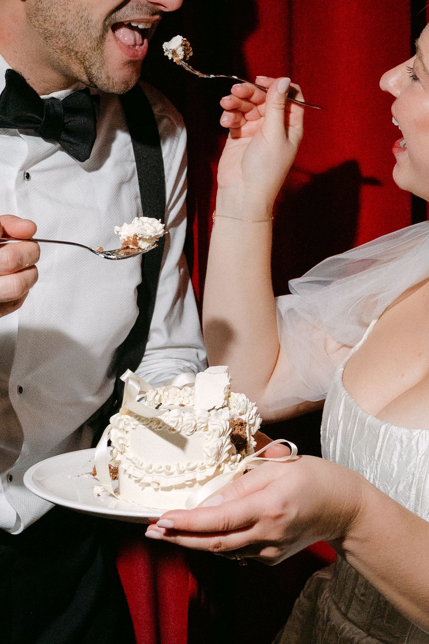 Newlyweds sharing wedding cake in a playful moment against a red backdrop.