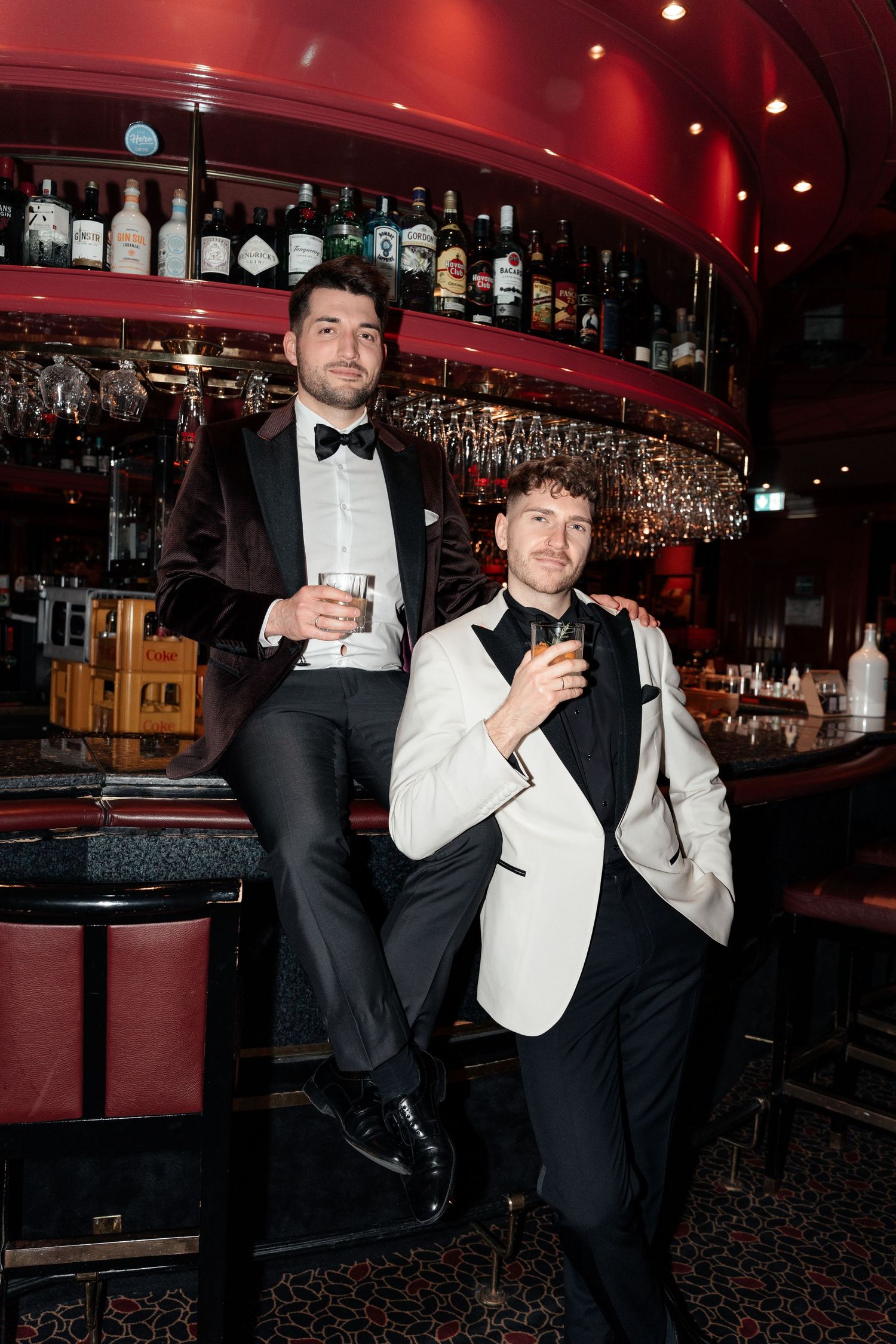 Two people in formal attire pose at an upscale bar with red decor and shelves of liquor bottles in the background.