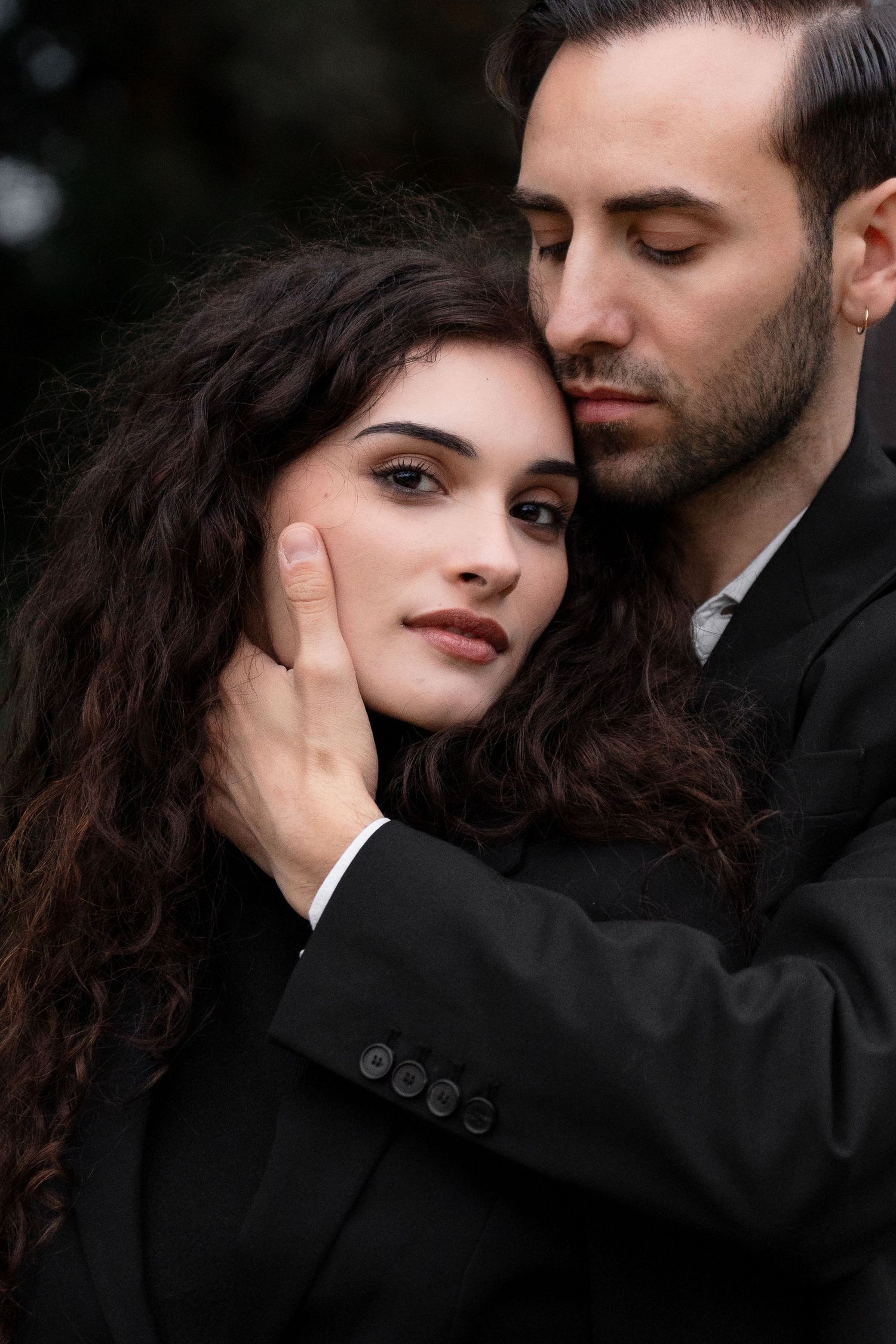 An intimate portrait of a couple in black clothing sharing a tender moment against a dark background.