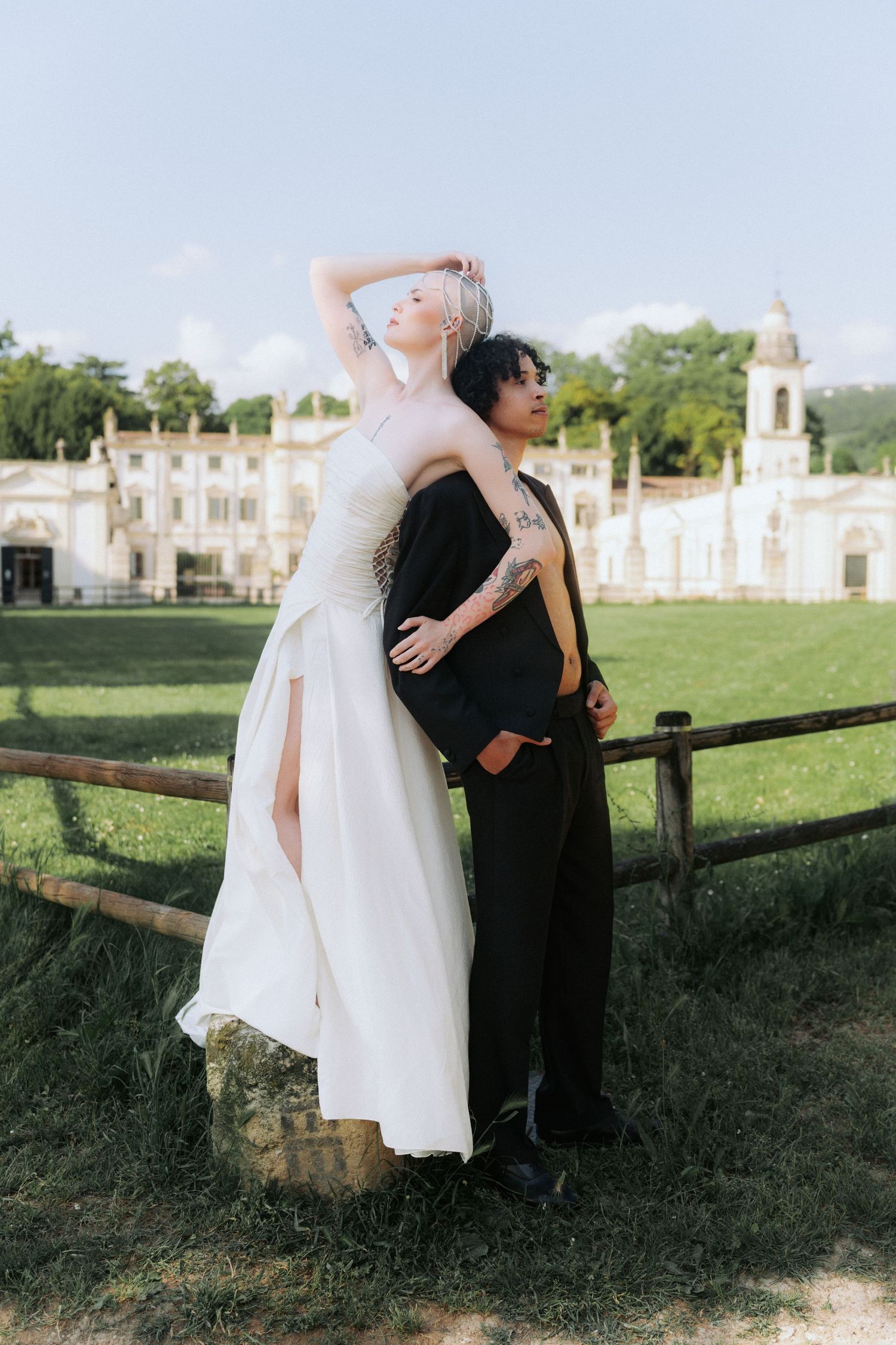 A couple in wedding attire poses against a rustic fence with an elegant historic estate building in the background.