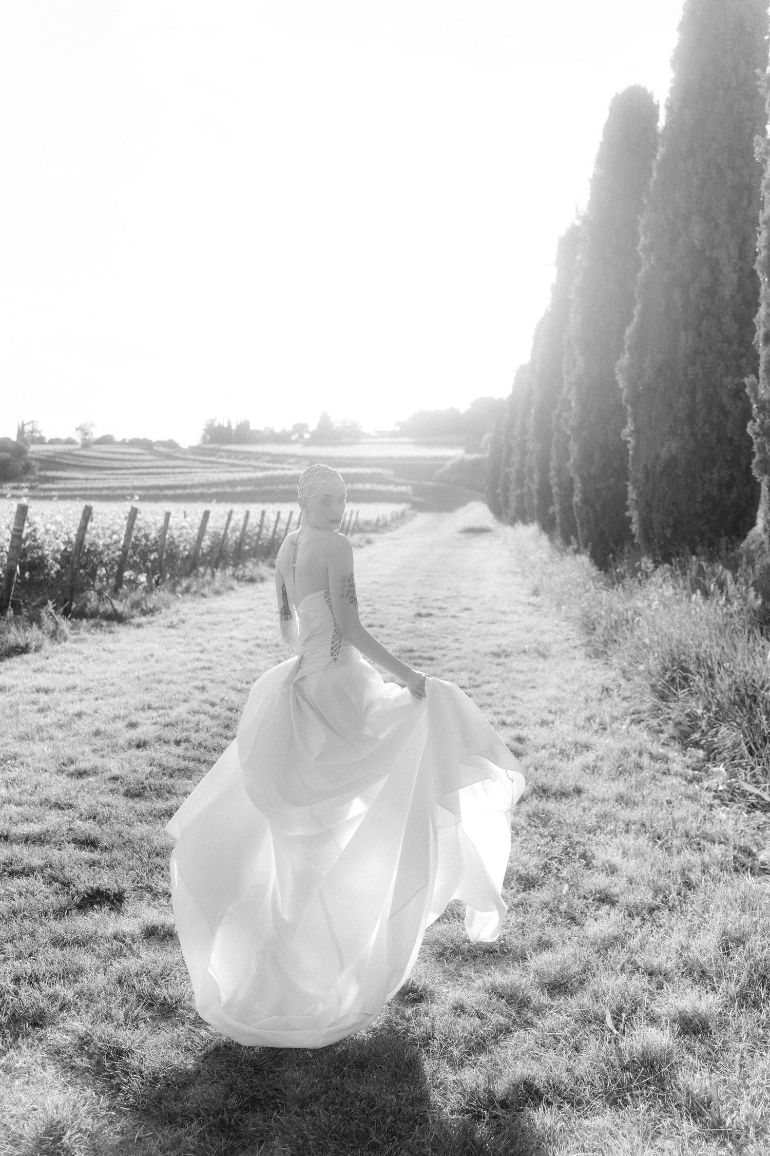 A dreamy black and white photo series shows a flowing white dress twirling in a sunlit field with cypress trees.