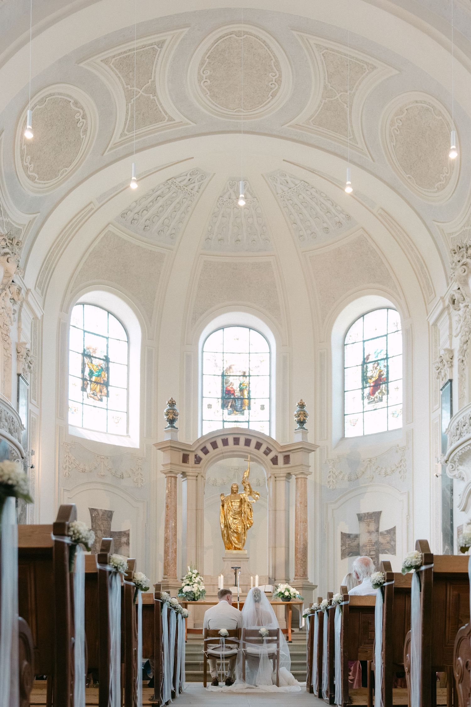 Interior view of an ornate white baroque church with vaulted ceilings, arched windows, and wooden pews.