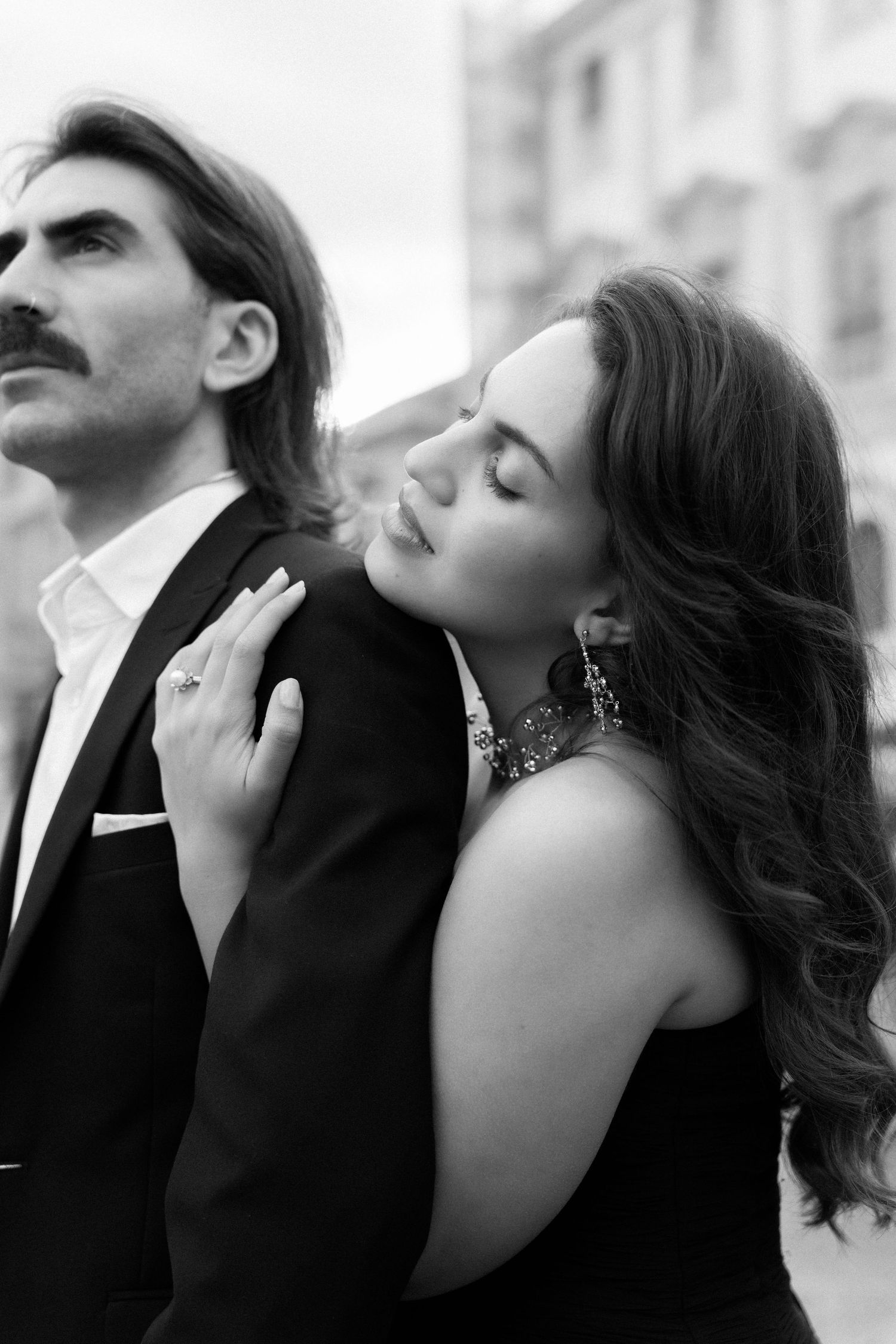 Black and white photo of an elegant couple, woman in black dress leaning on mans shoulder in a romantic urban setting.
